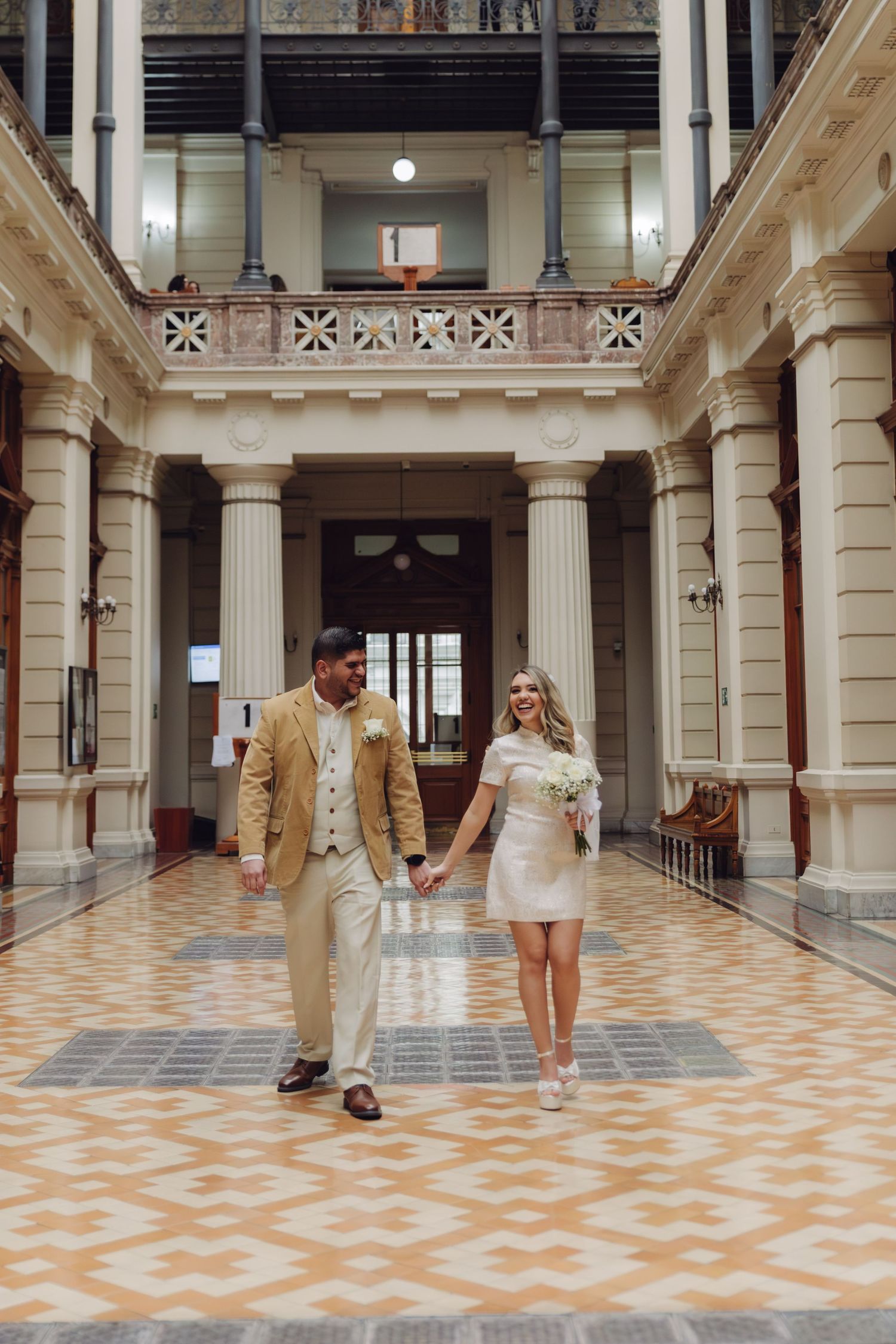 Smiling bride and groom in casual wedding attire walking through a beautiful historic building interior.