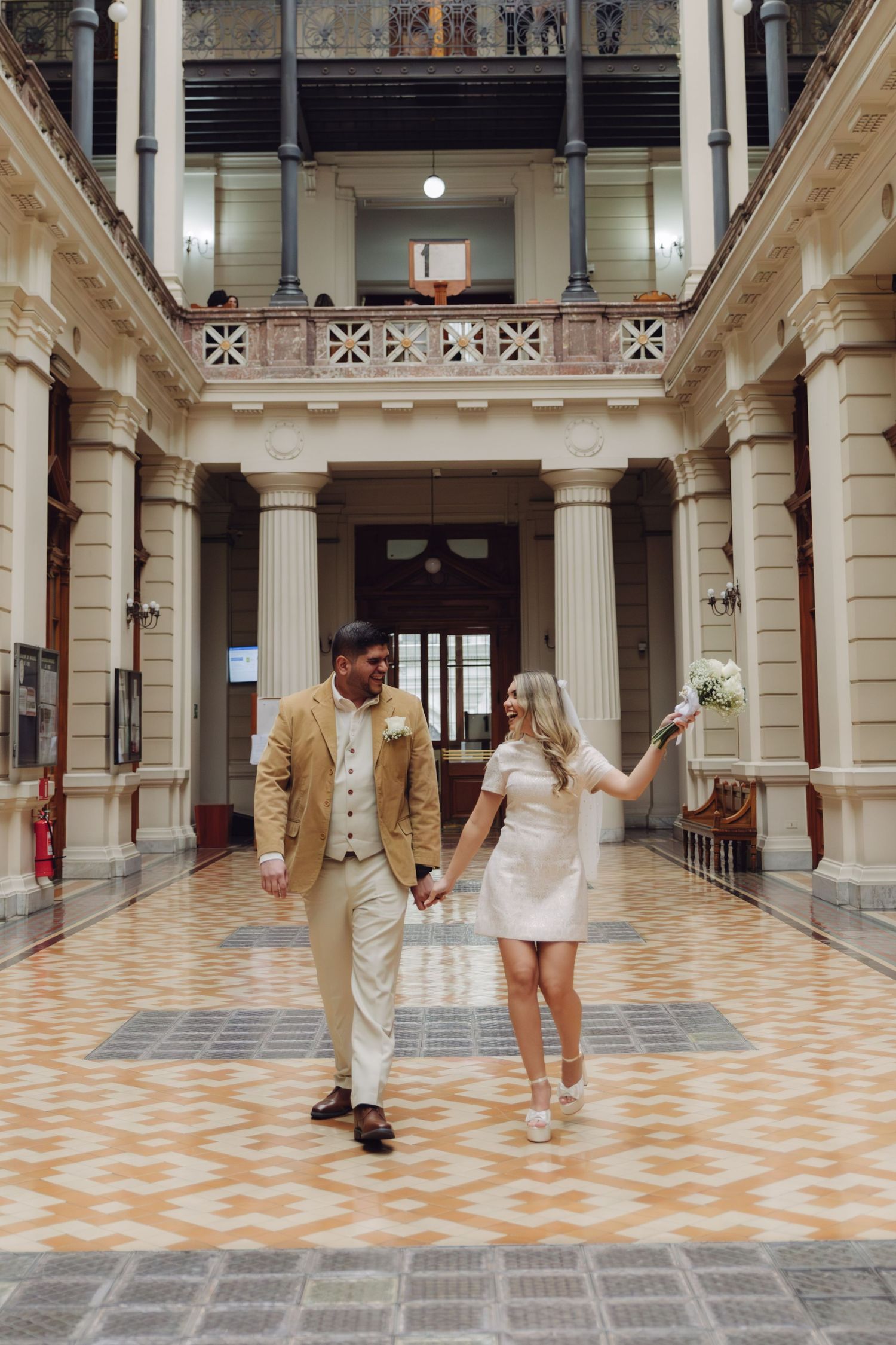 Joyful couple in wedding attire walking through ornate hall, bride raising white bouquet while holding grooms hand.