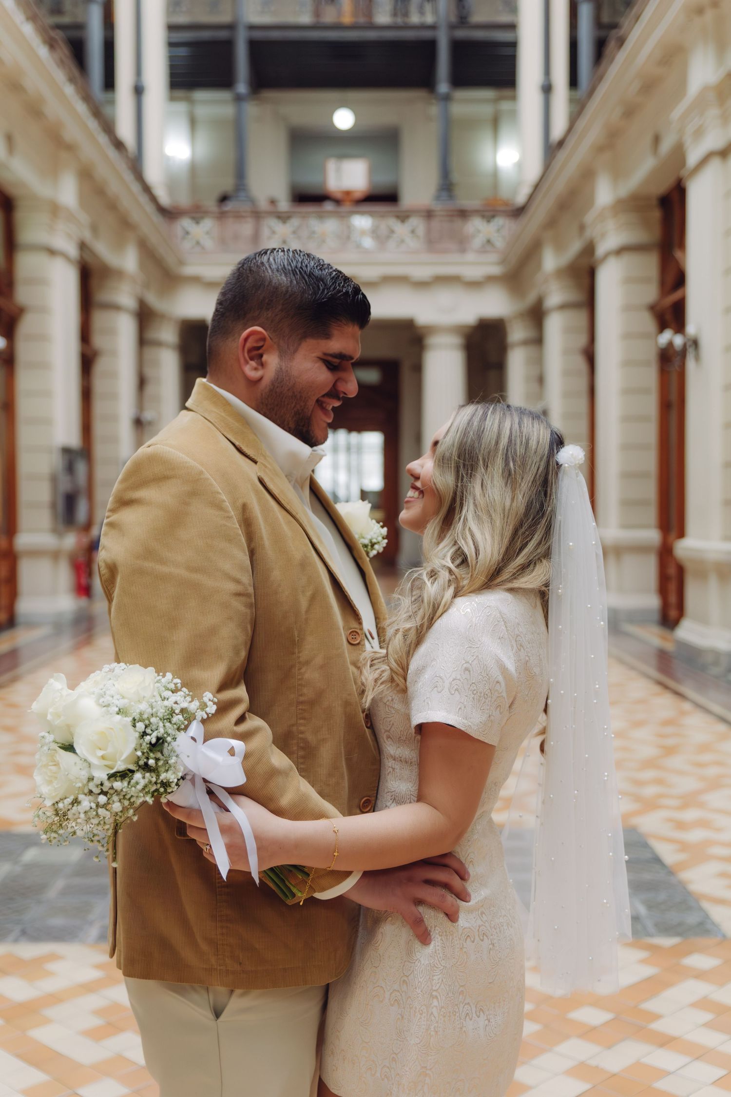 Bride and groom embracing and smiling at each other in a grand historic venue with white columns.