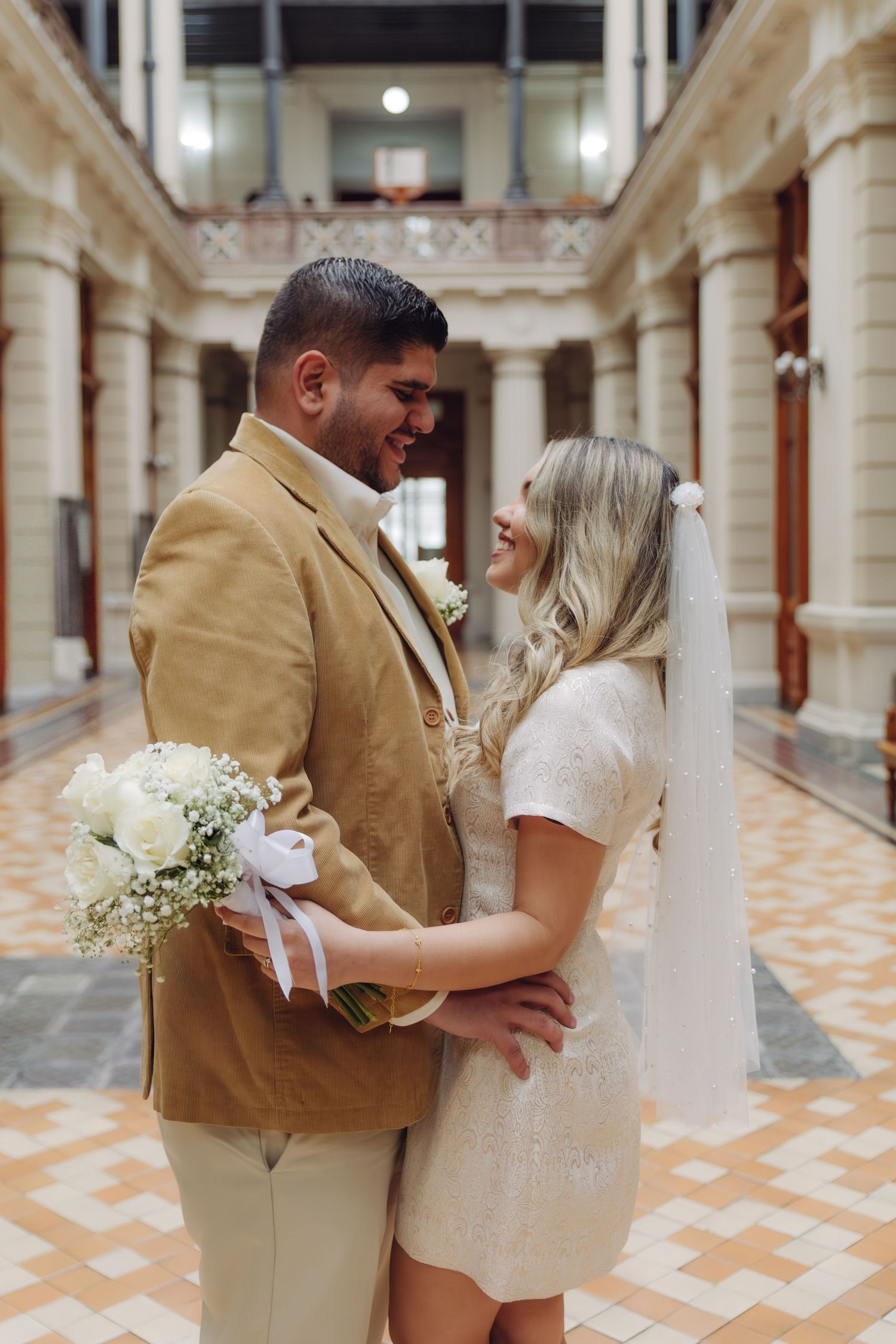 Newlywed couple sharing a romantic moment in a stunning colonial building, groom holding white rose bouquet.