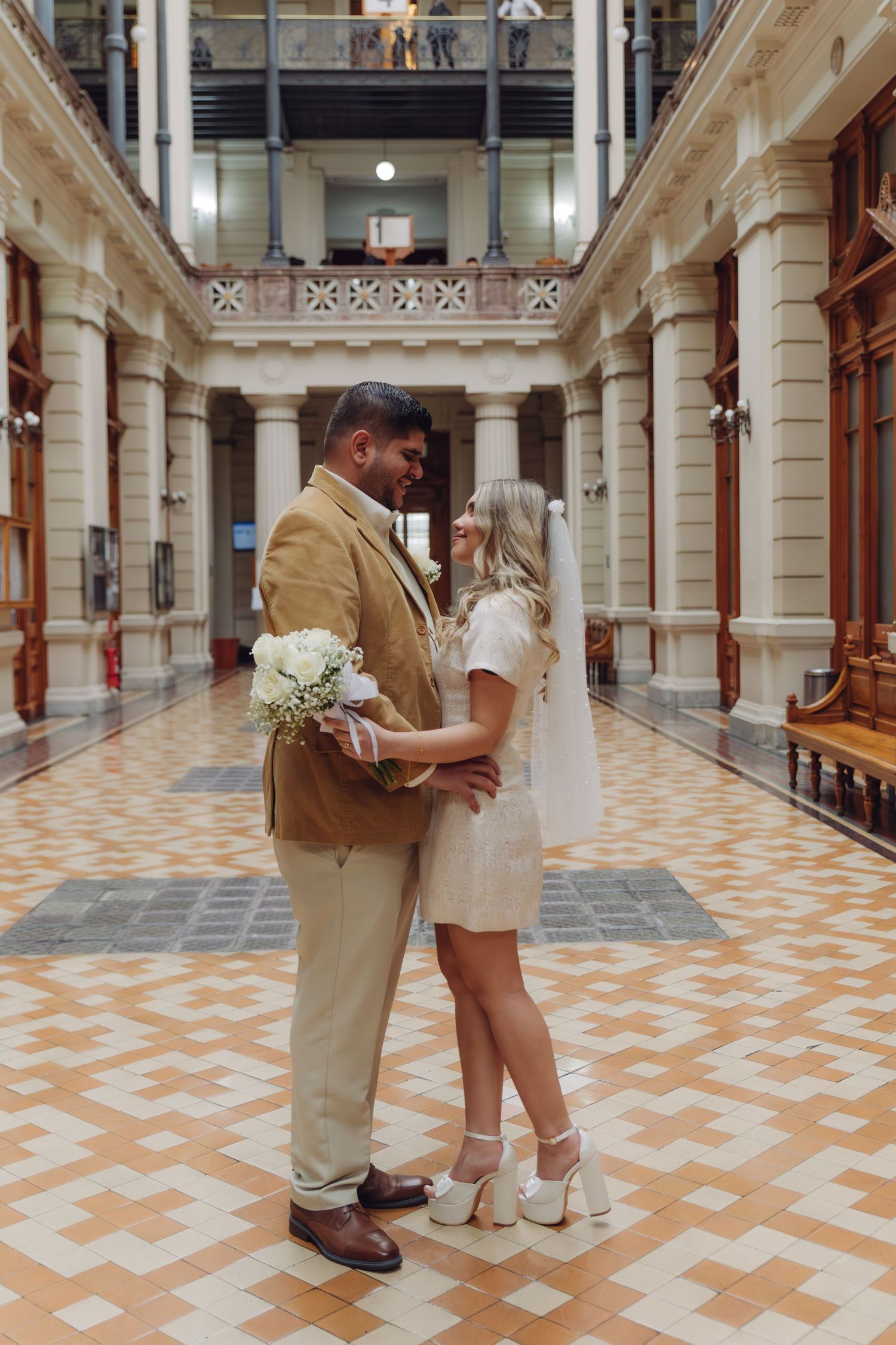 Elegant wedding couple standing close together in a grand historic hall with tall decorative columns.
