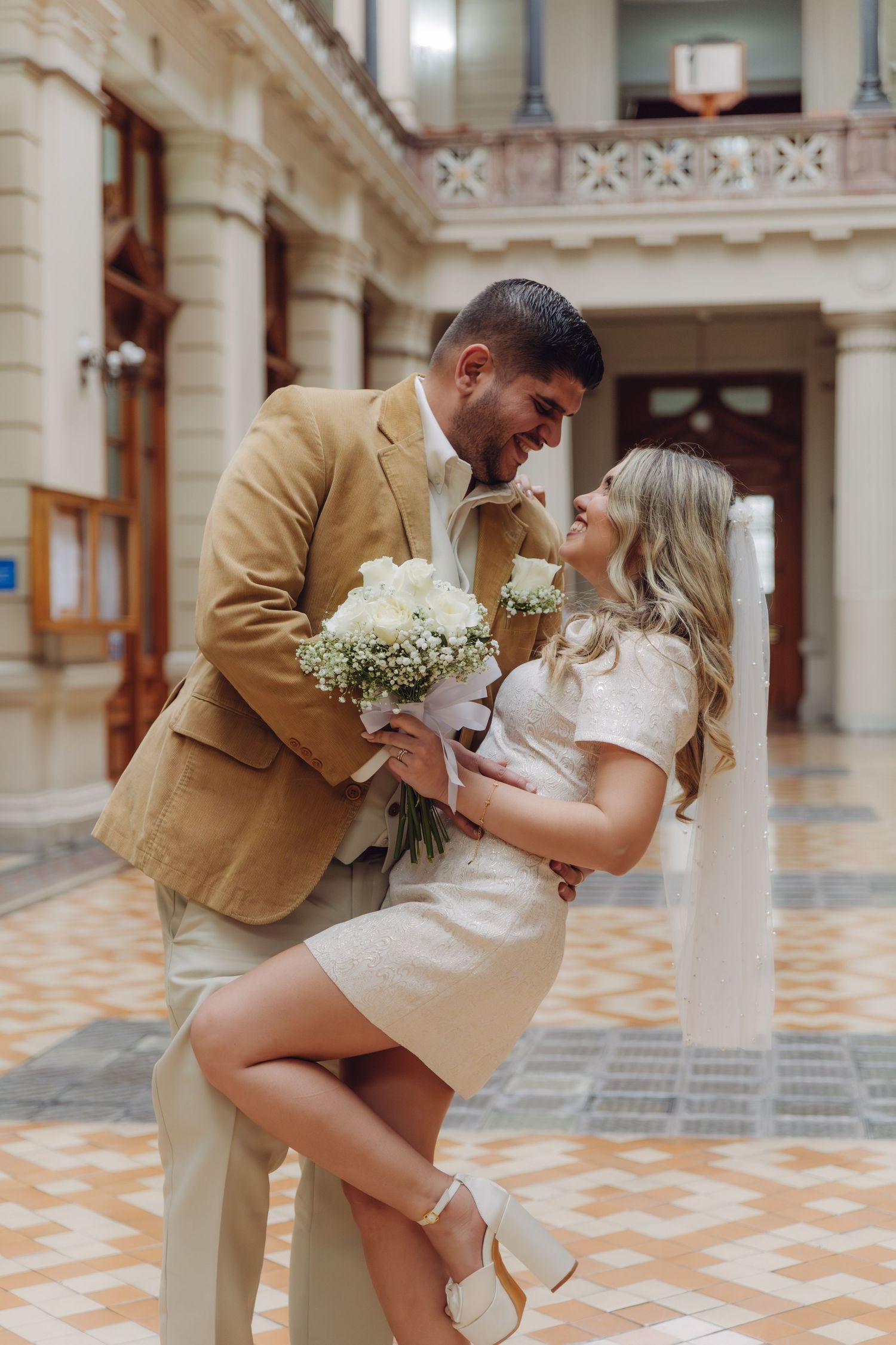 Couple sharing romantic moment, groom in tan suit holding white bouquet, bride in short white dress seated, elegant indoor venue.