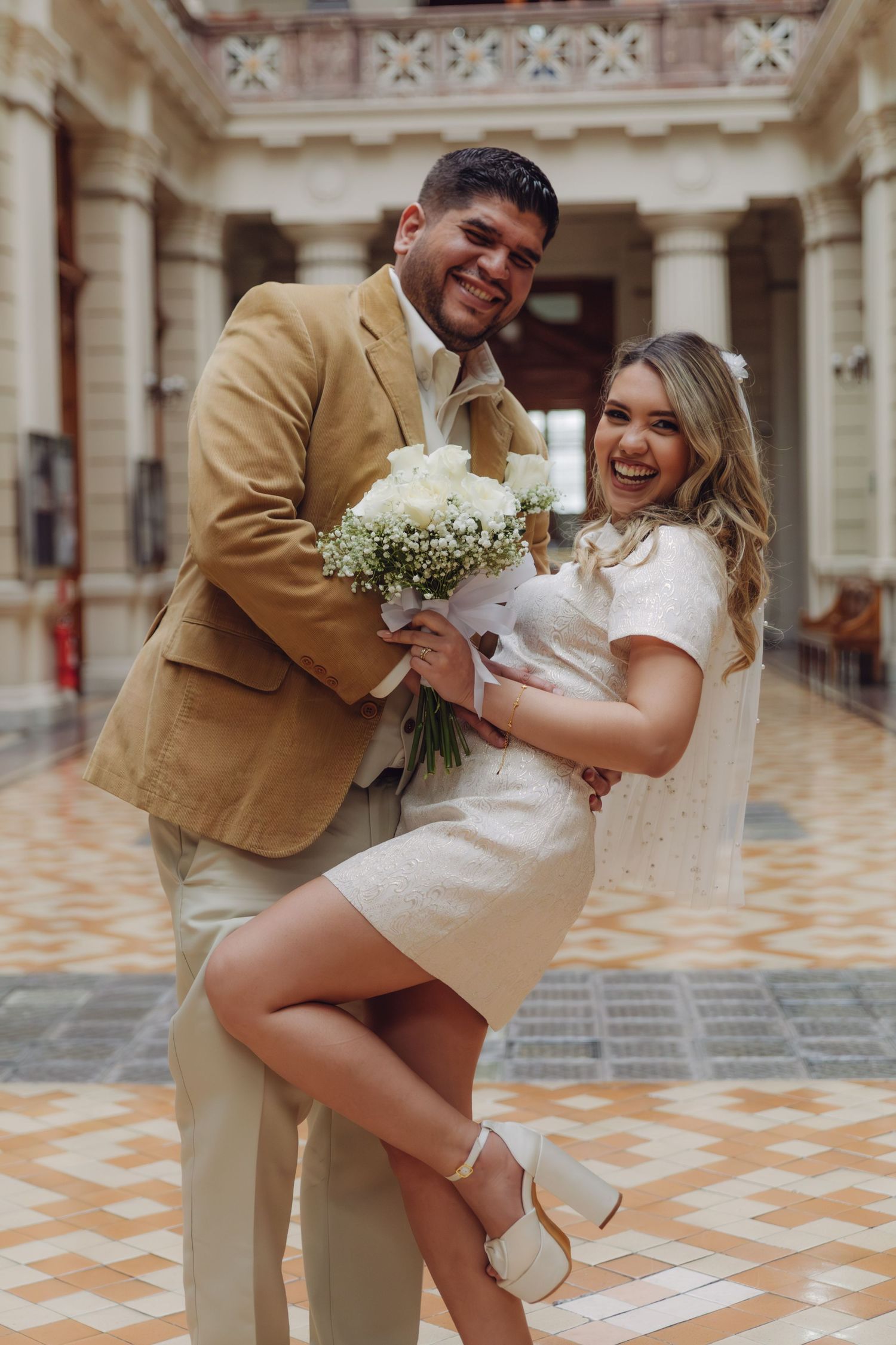 Joyful newlyweds laughing together indoors, groom in tan blazer, bride in white mini dress holding white floral bouquet.