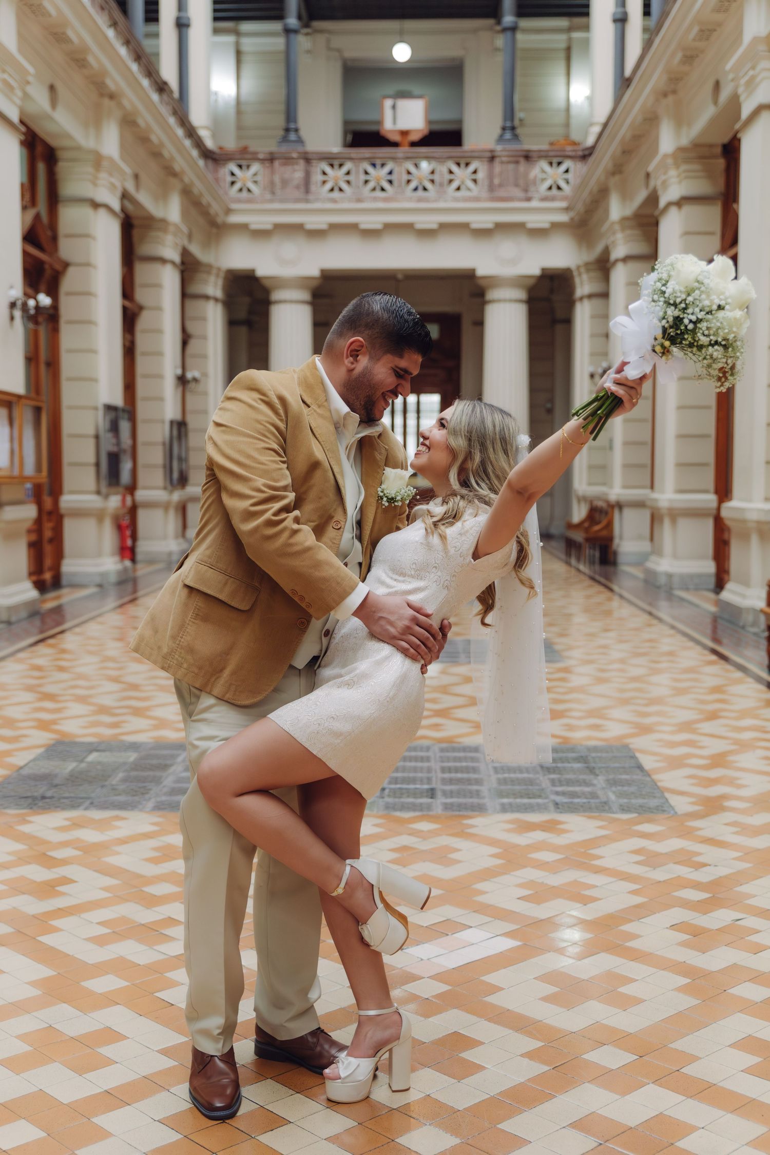 Bride and groom sharing a dip kiss, bride raising white bouquet, stunning tiled floor in grand historic building.