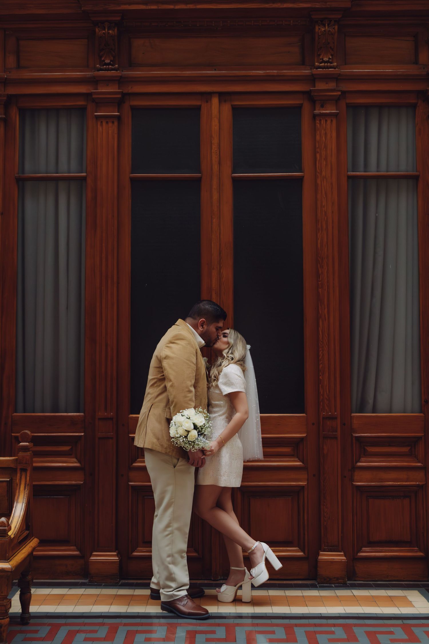 Newlyweds sharing a kiss in front of ornate wooden doors, bride in short white dress, groom in tan suit.