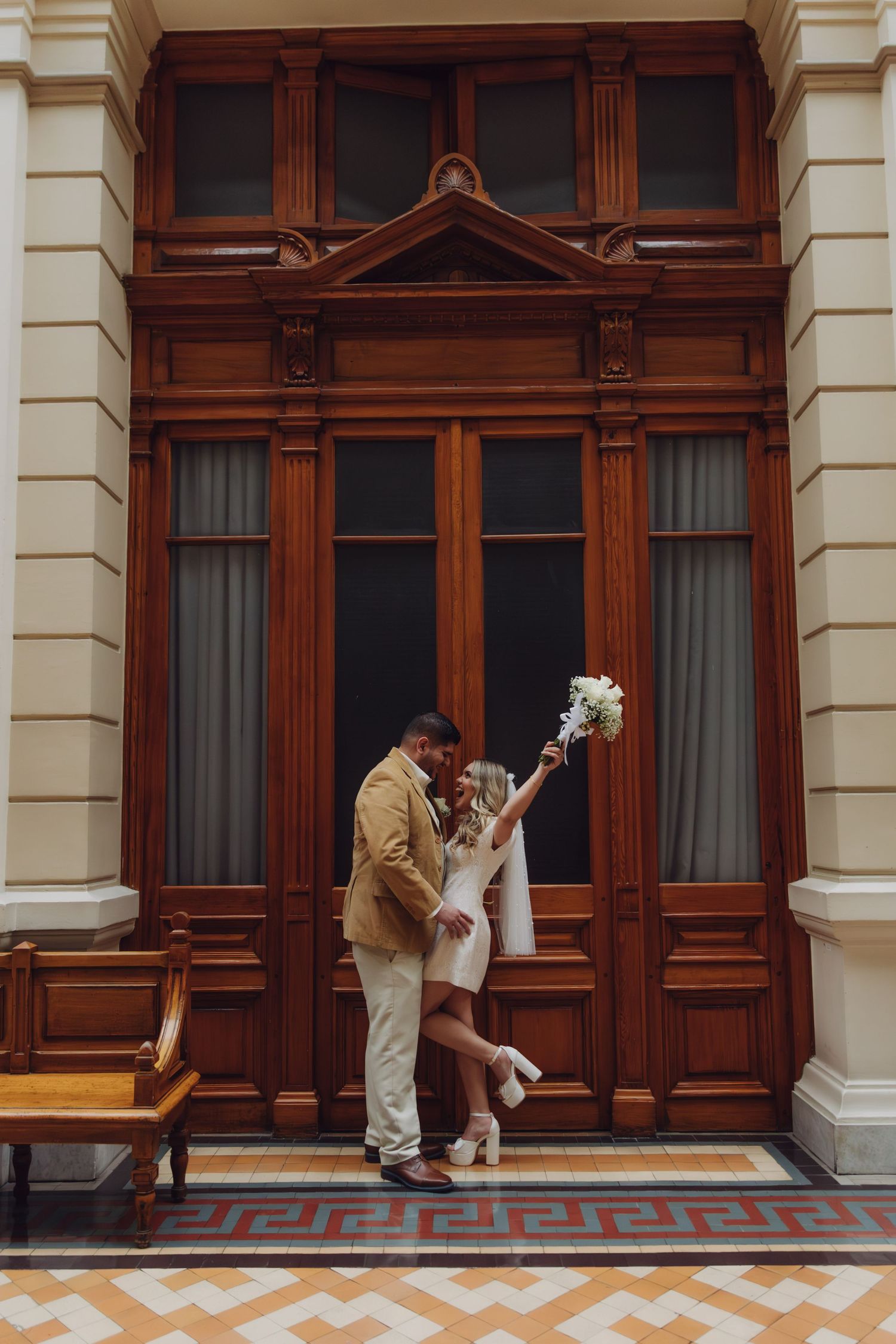 Couple embracing before grand wooden doors, bride in white mini dress raising bouquet, groom in tan blazer outdoors.