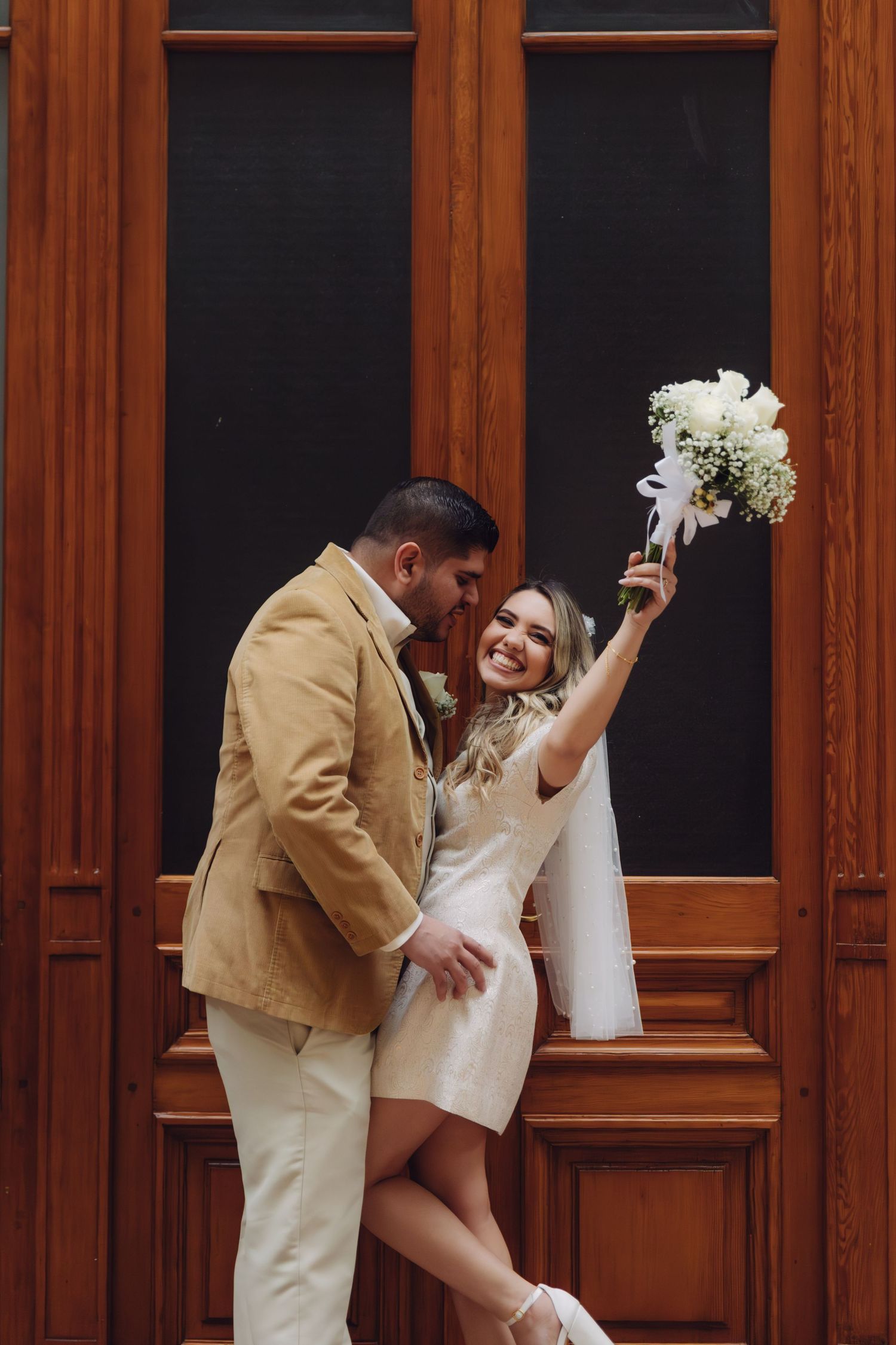 Smiling bride raising white bouquet beside groom in tan jacket, posing in front of elegant wooden doors.