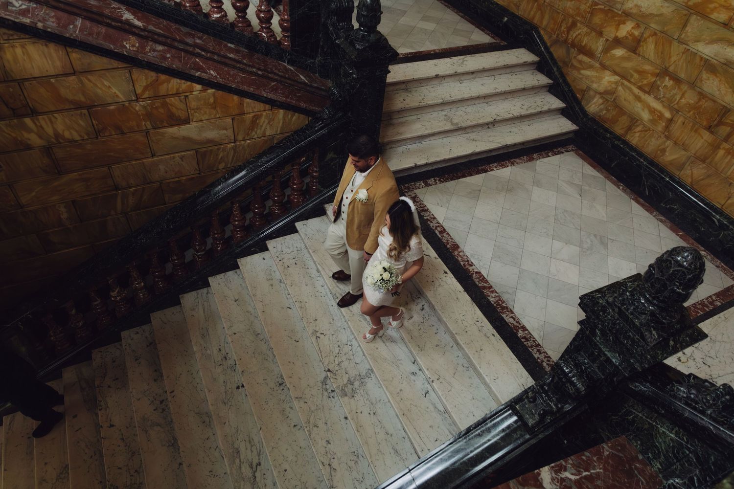 Aerial view of bride and groom descending a grand marble staircase with ornate dark railings in historic building.