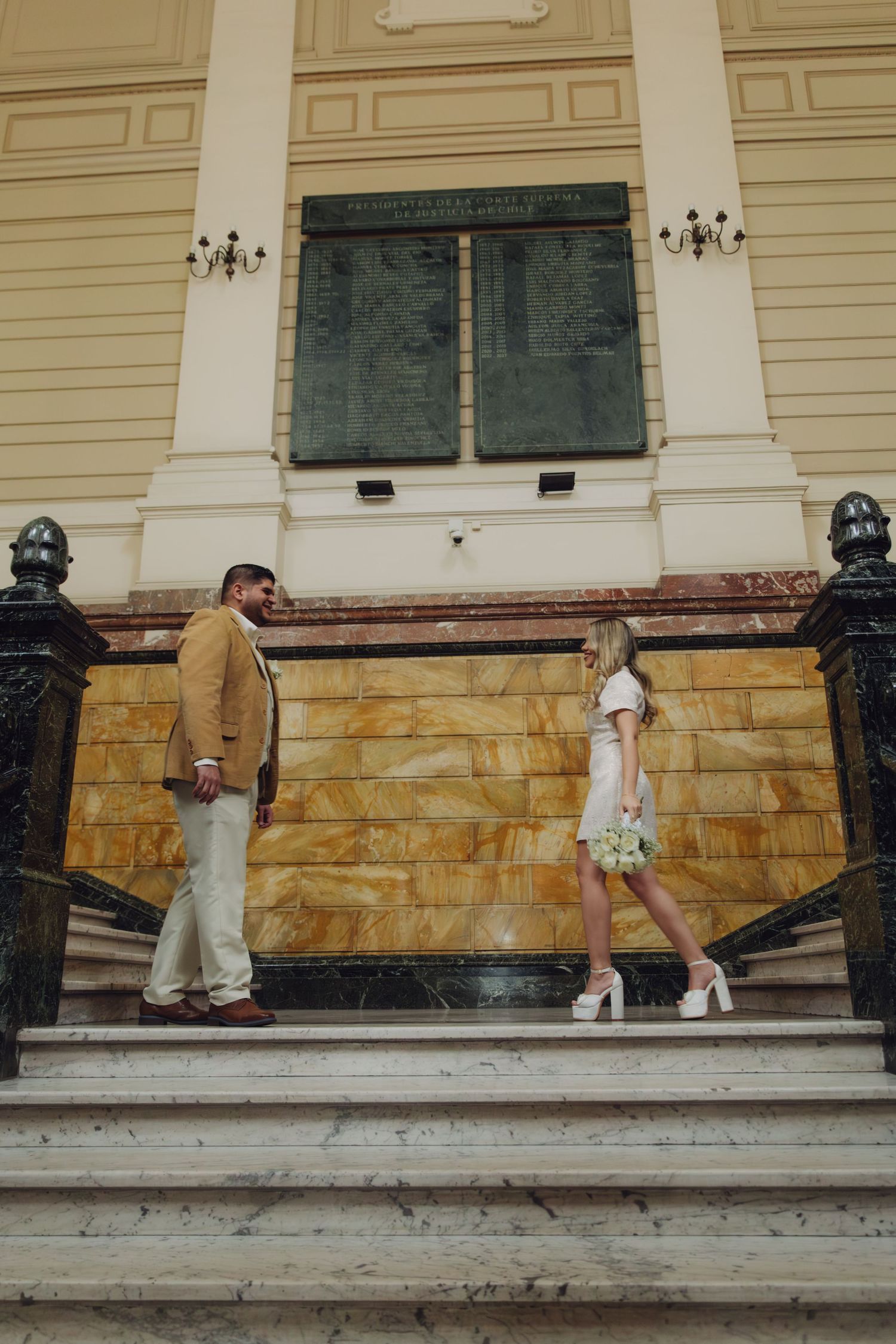 Stylish couple stands on marble steps of grand classical yellow building, bride holding white bouquet in mini dress.