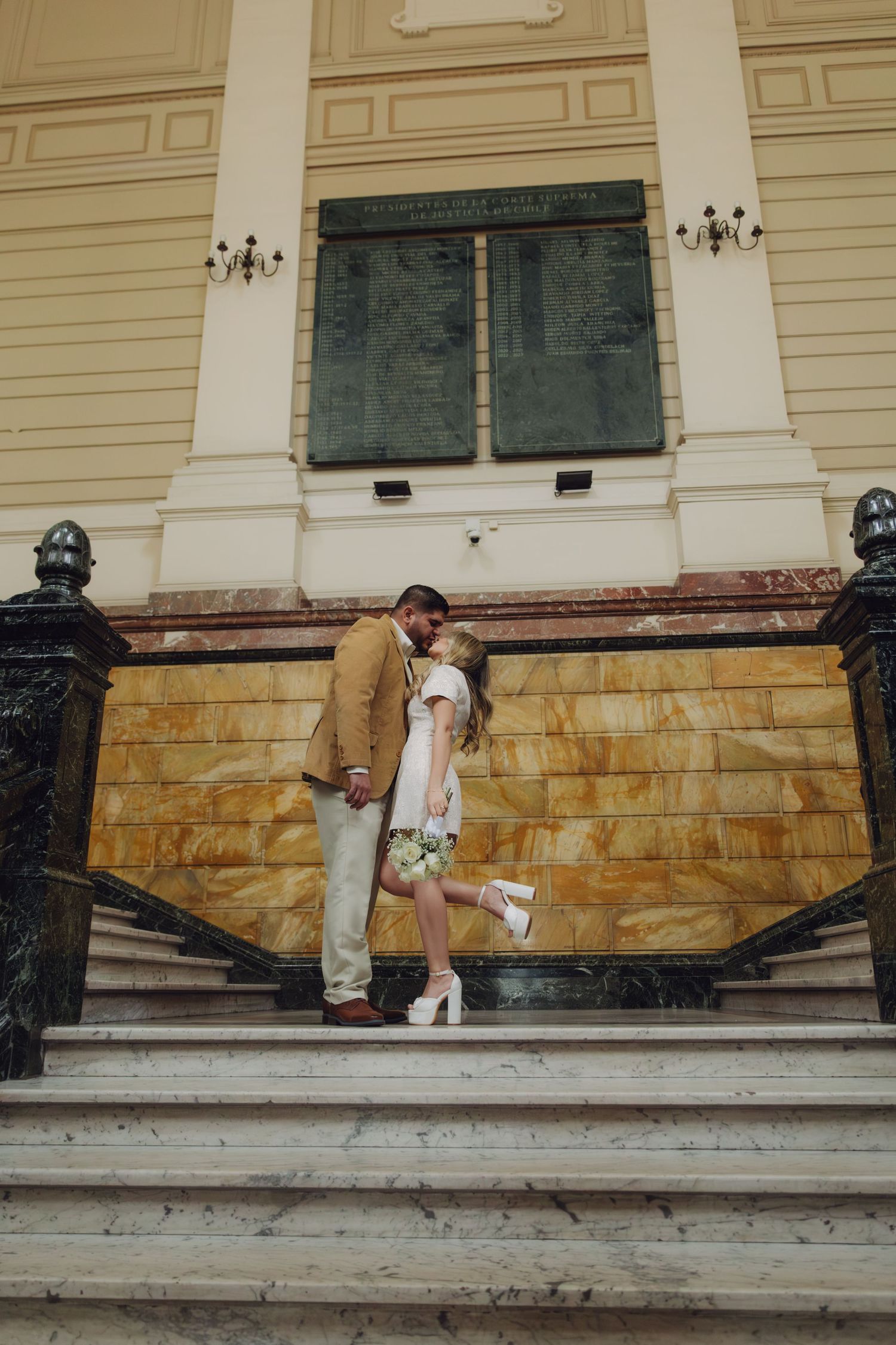 Couple shares romantic kiss on grand marble staircase outside ornate yellow classical building with decorative columns.