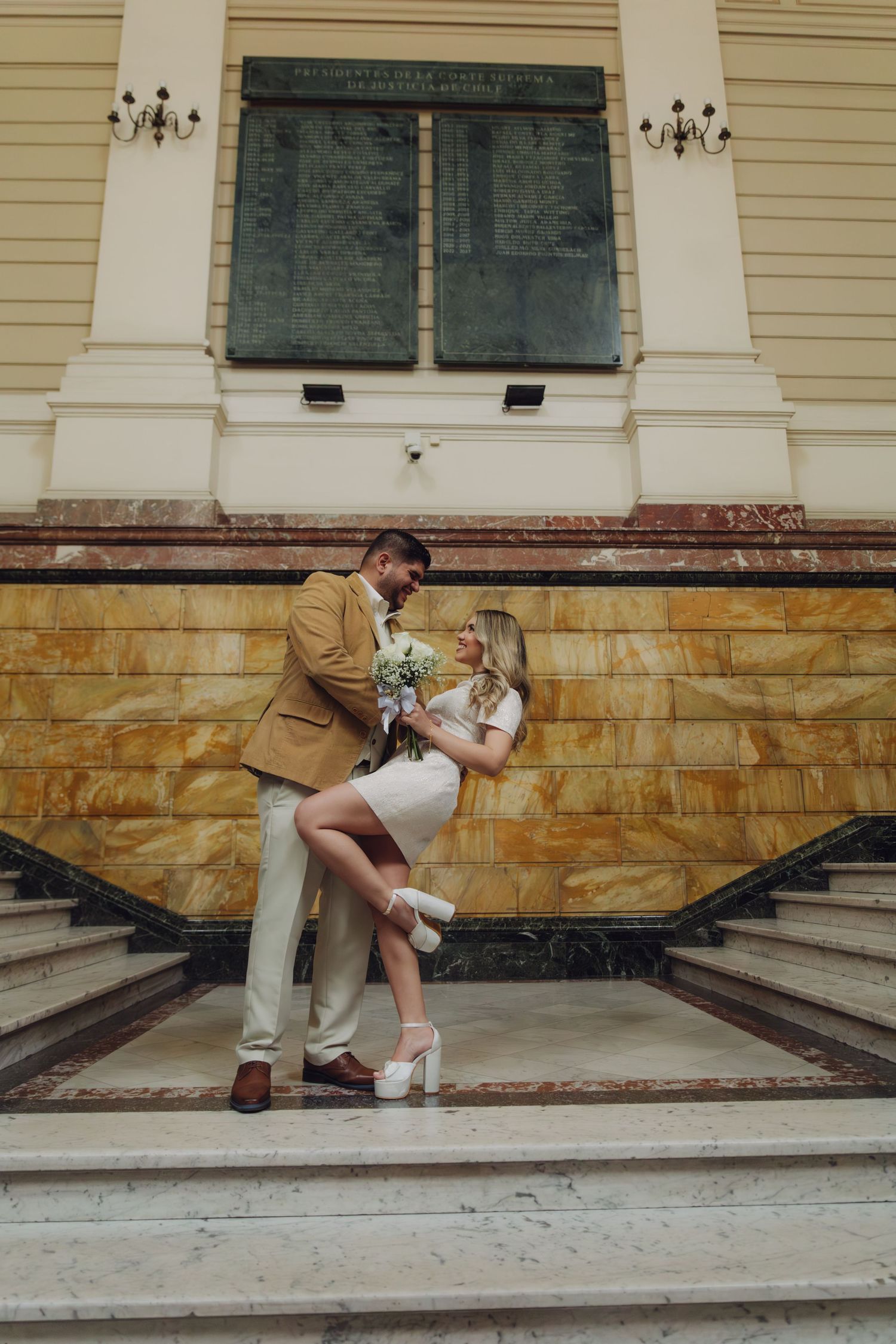 Couple embracing on marble steps of elegant building, groom in tan blazer, bride in white mini dress holding white bouquet.