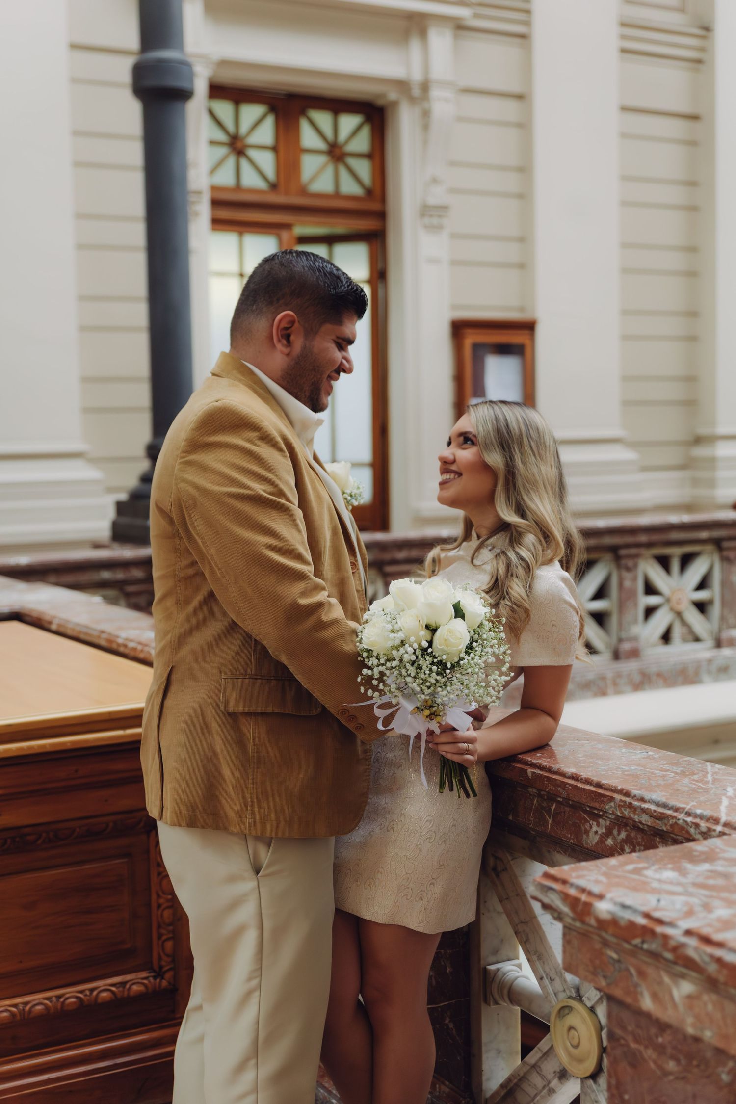 Newlyweds sharing romantic moment on ornate marble balcony, bride holding white rose bouquet, groom in camel blazer.