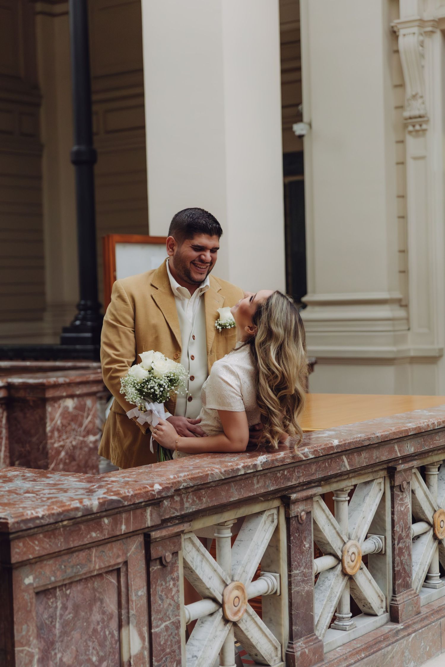 Smiling couple embracing on decorative marble railing, groom in tan jacket, bride with flowing blonde hair and white bouquet.