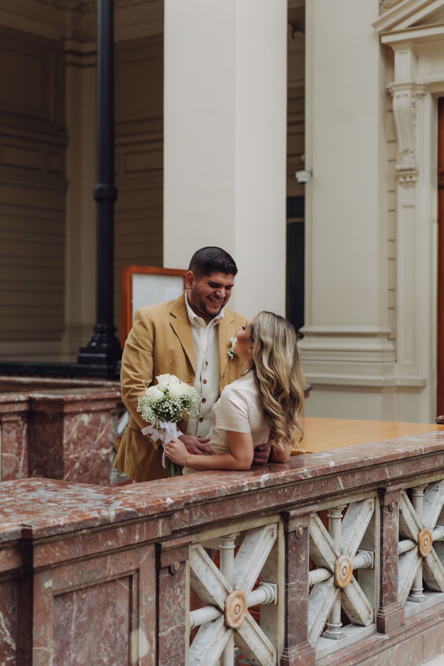 Wedding couple posing on ornate marble balcony with carved railings, groom in camel blazer holding bride with white flowers.