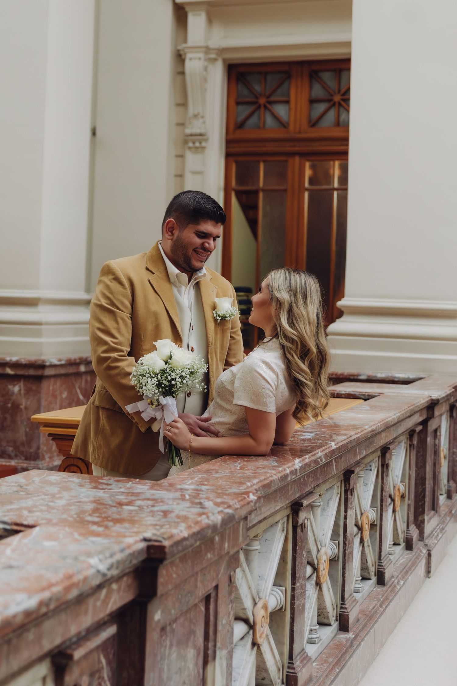 Elegant couple sharing intimate moment on marble balcony, bride in white dress holding bouquet, groom in tan blazer.