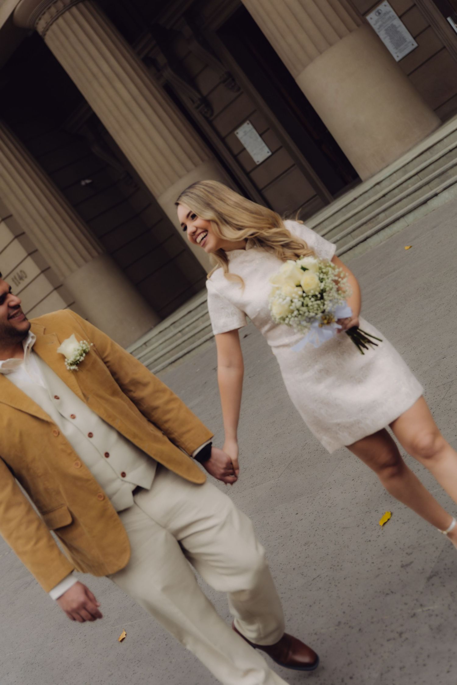 Joyful bride in white mini dress holding bouquet walking with groom in tan blazer outside grand columned building.