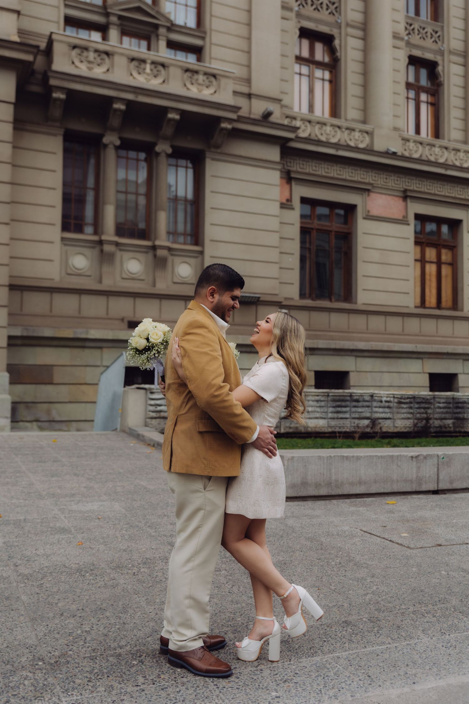 Wedding couple embracing and smiling outside historic European building, bride in white dress, groom in camel blazer.