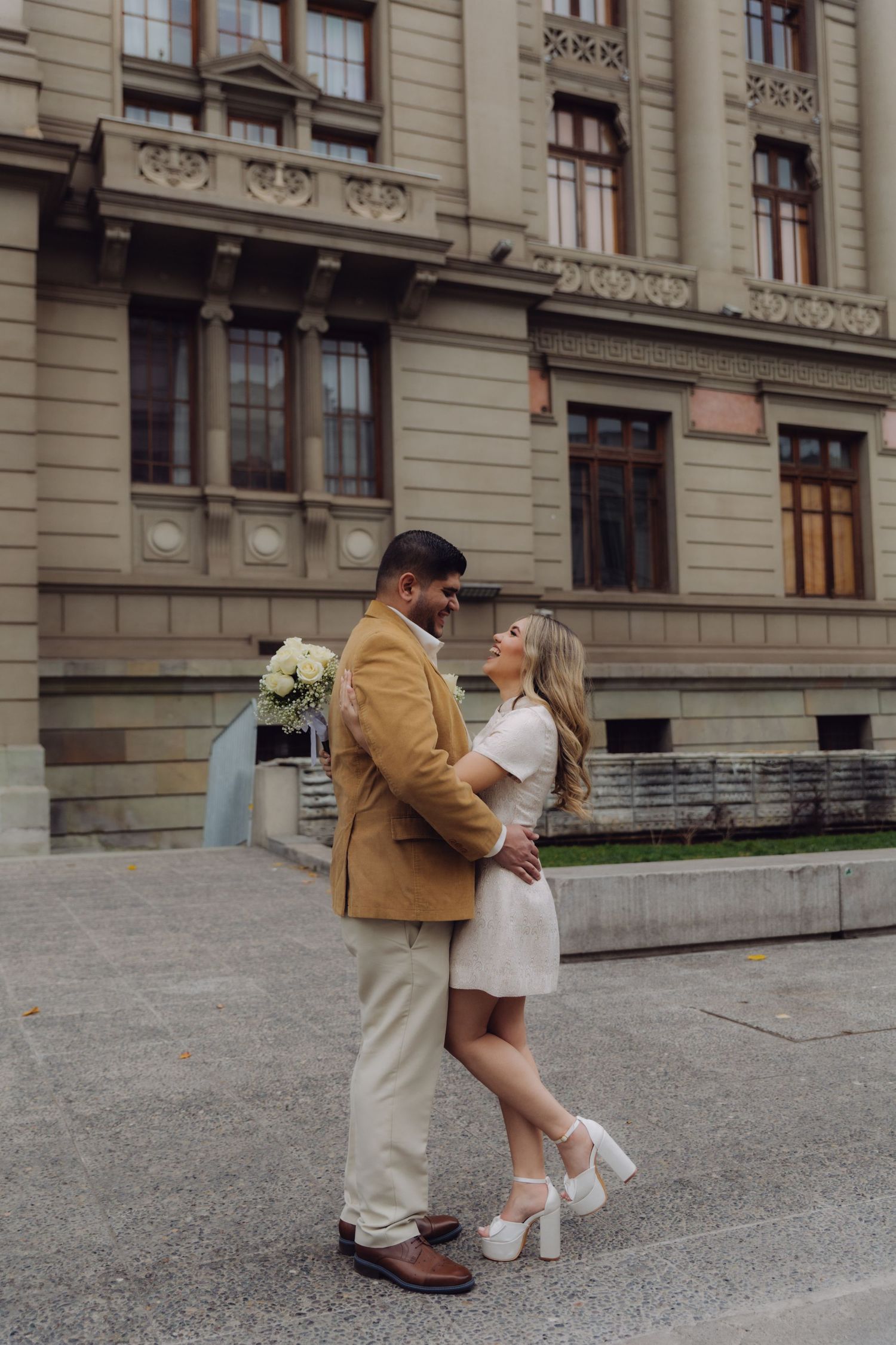 Newlywed couple sharing romantic embrace outside grand historic building, bride in white dress, groom holding white bouquet.