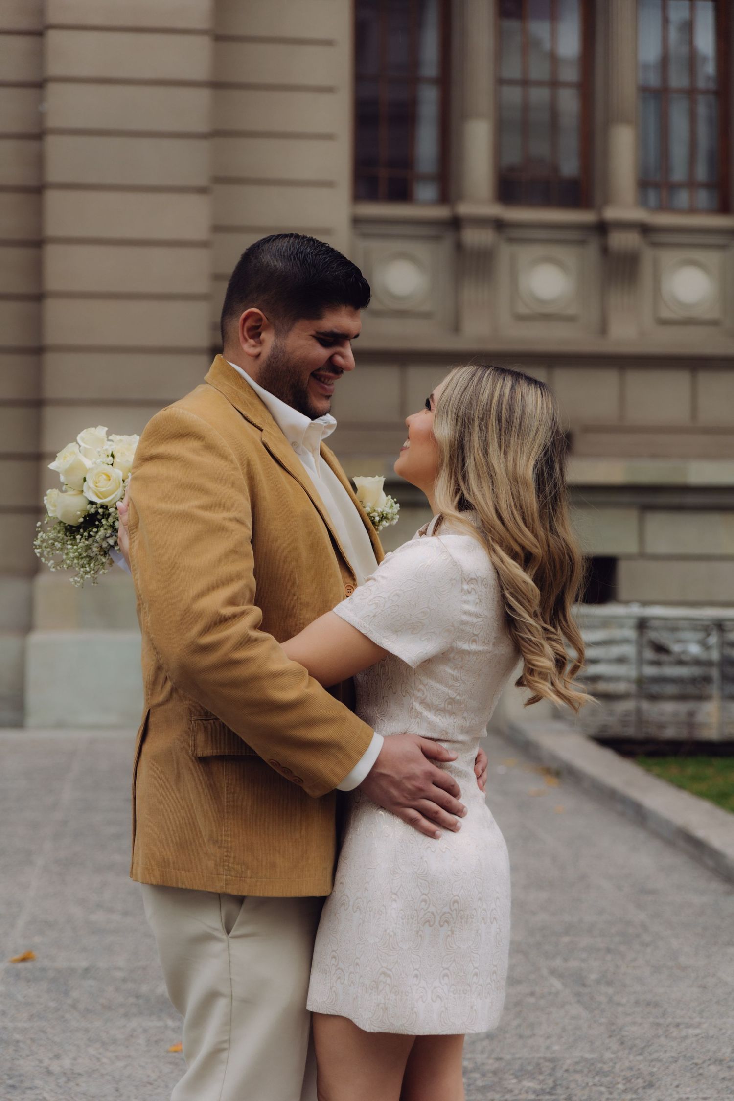 Groom in tan blazer embracing bride in white textured dress outdoors, holding white rose bouquet near historic building.