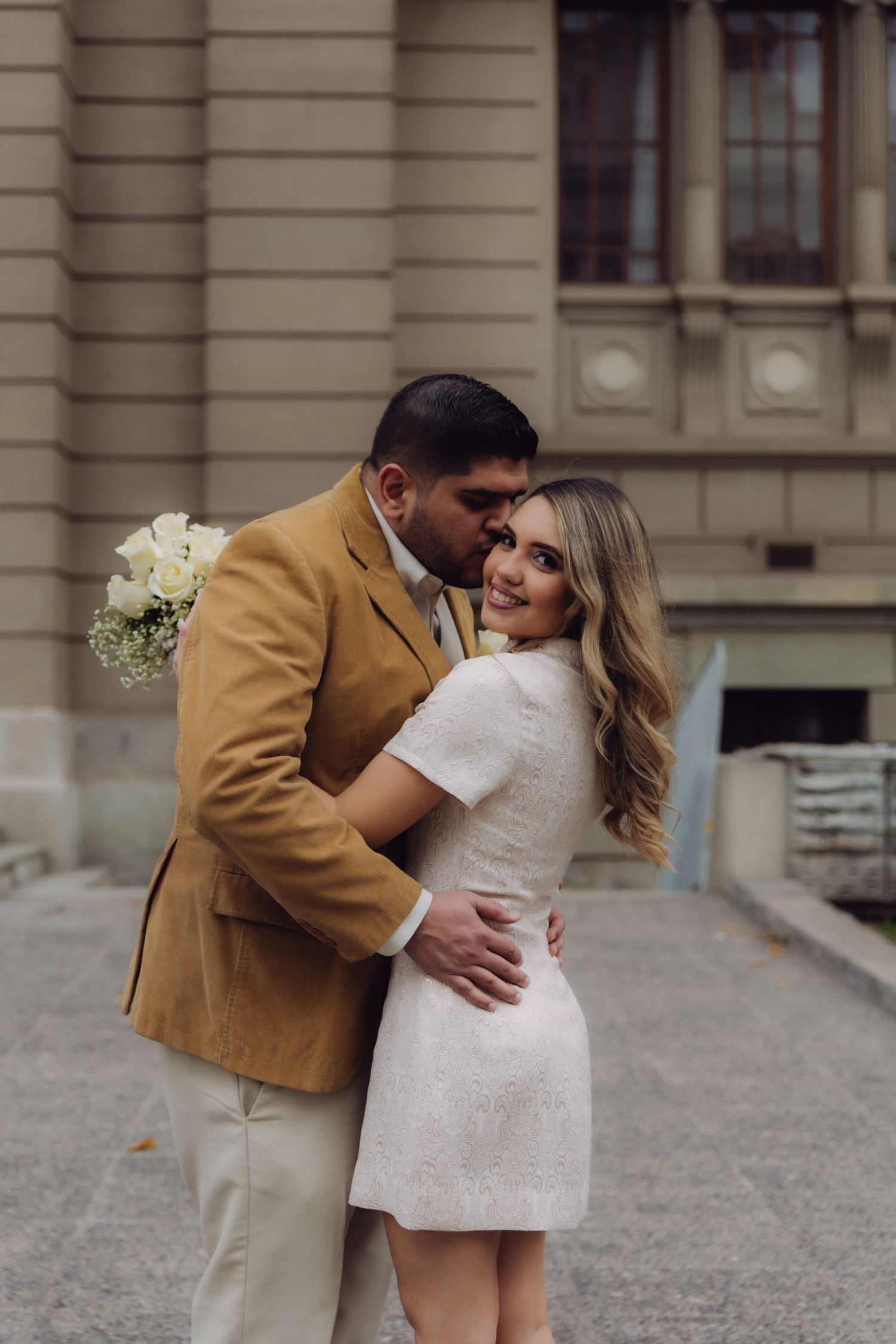 Groom kissing bride on temple outdoors, bride smiling at camera in white mini dress, groom in camel blazer with bouquet.