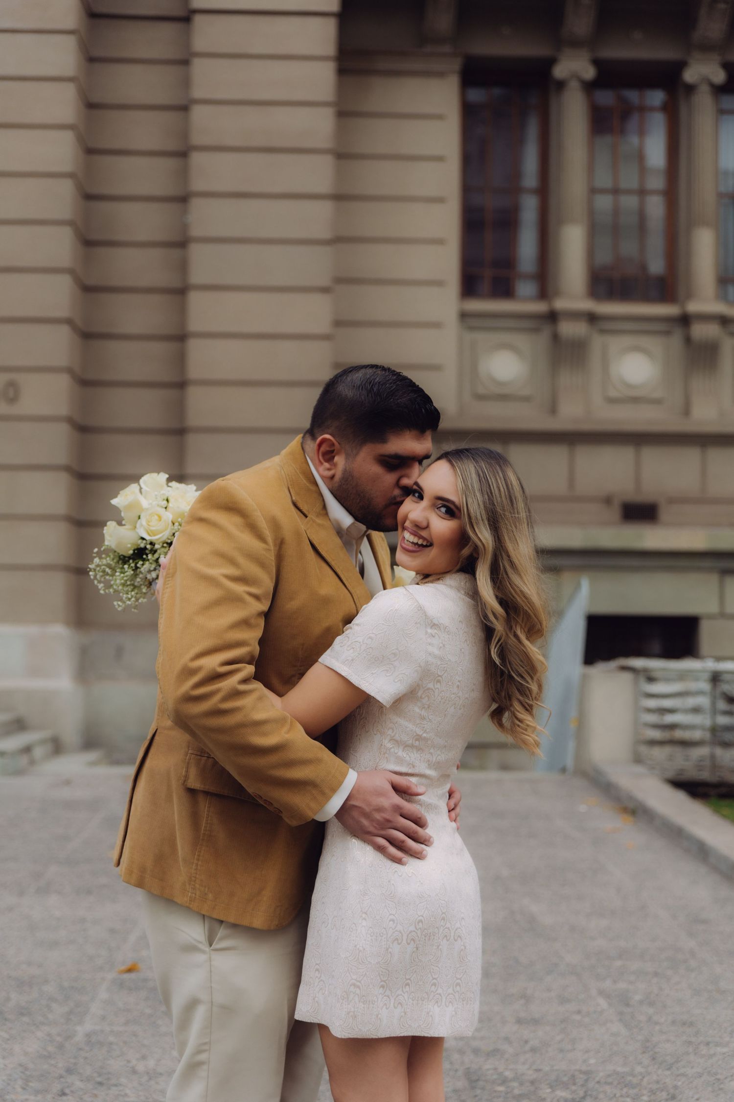 Couple embracing outside historic building, groom in tan suit kissing bride in white mini dress holding bouquet.