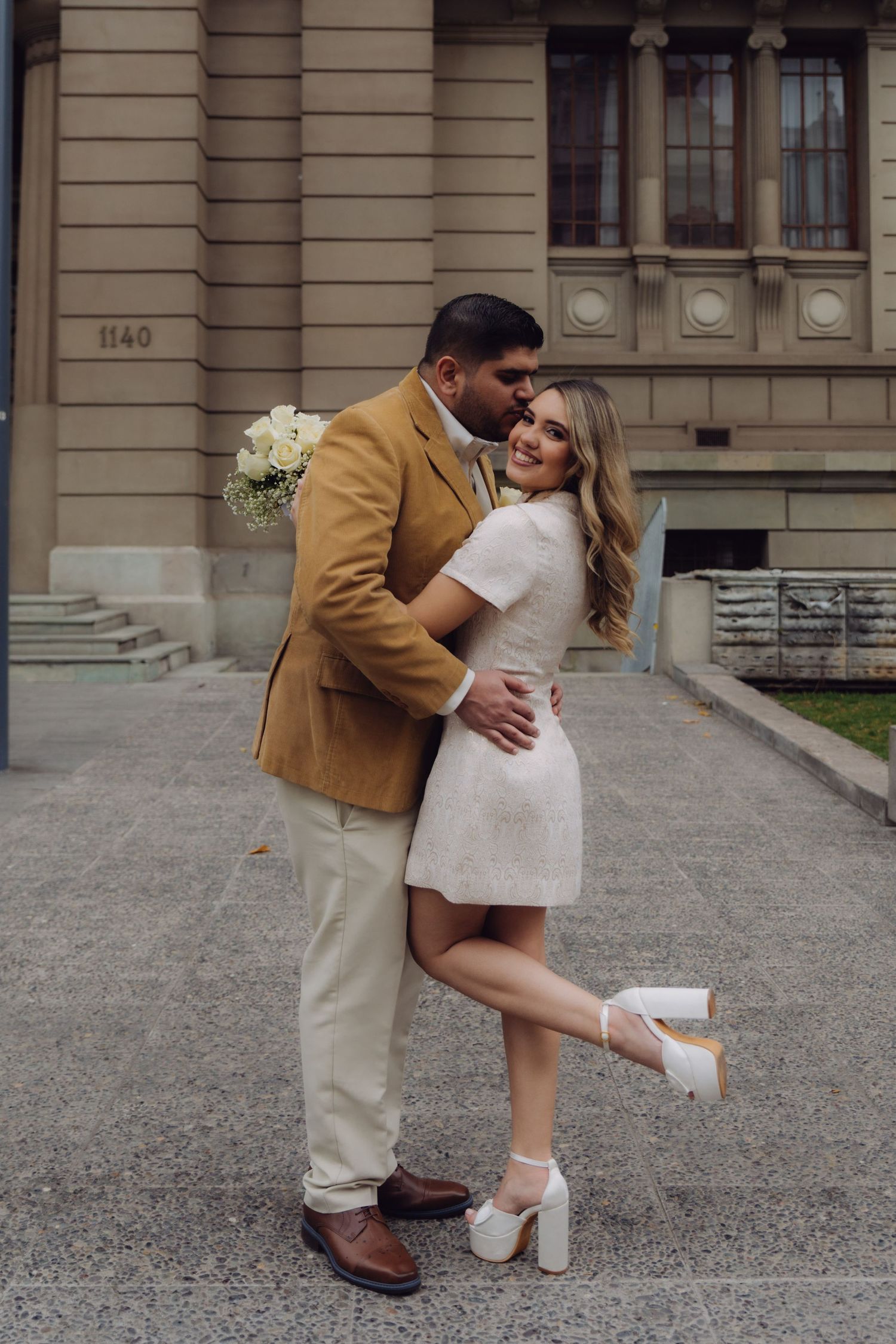 Bride in white mini dress and heels lifts leg as groom in tan suit embraces her outside stone building.