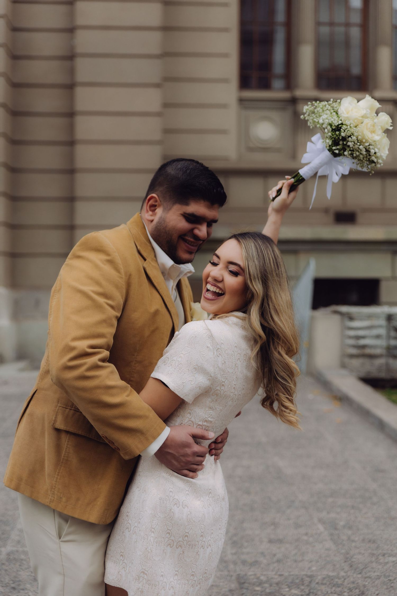 Laughing bride in white dress raises bouquet while groom in tan blazer hugs her outside historic building.