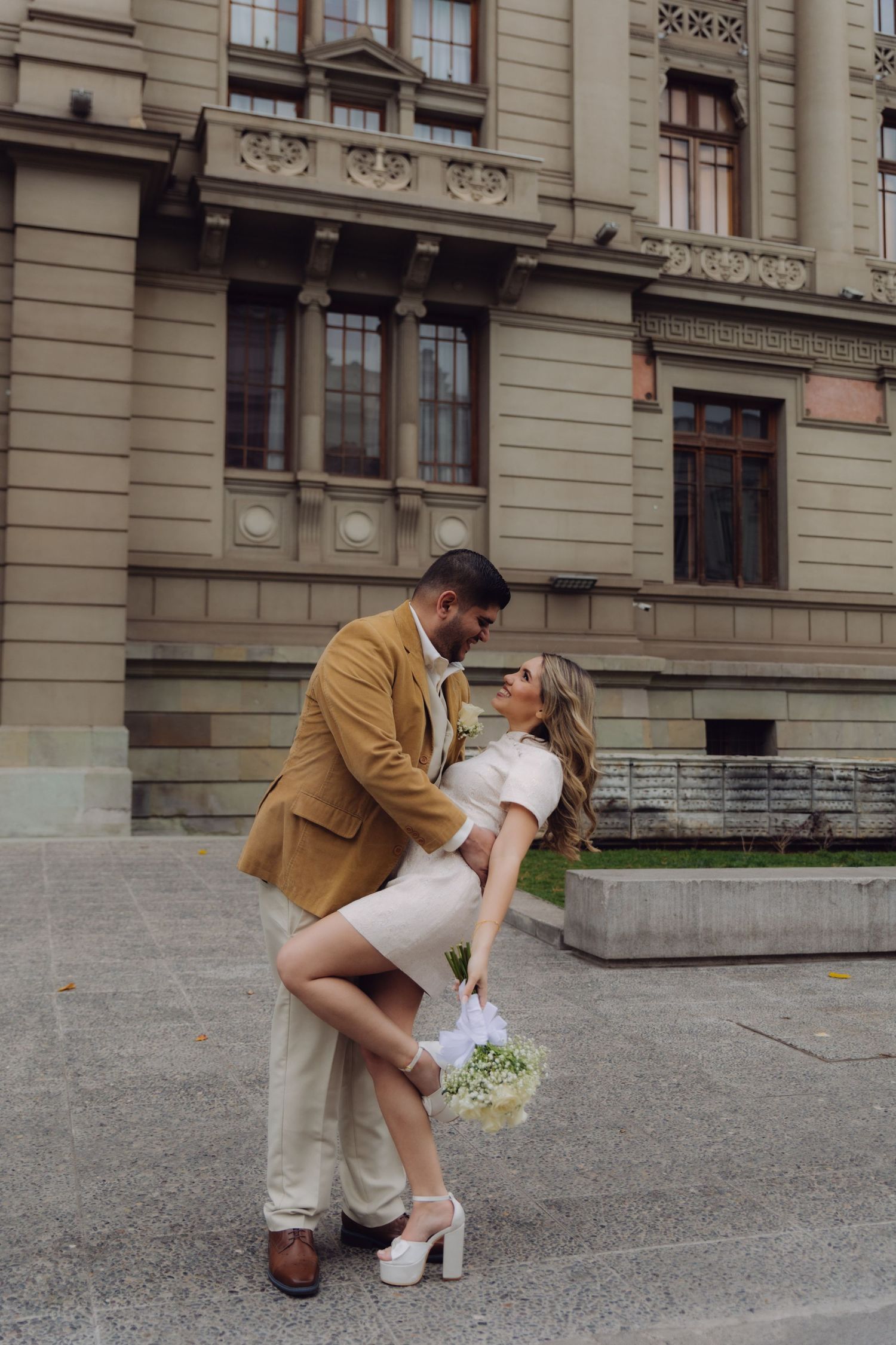 Groom in tan suit dips bride in white mini dress holding bouquet outside grand stone building.