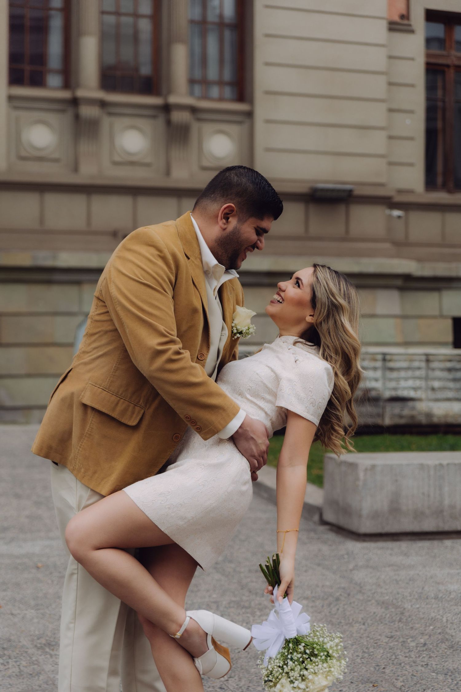 Smiling couple sharing romantic dip pose outside historic building, groom in tan suit and bride in white dress.