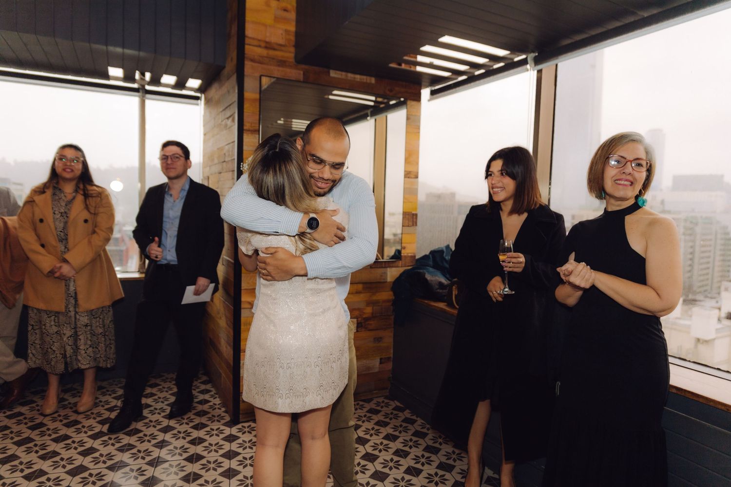 Newlyweds share a hug at rooftop reception while guests smile and celebrate around them.