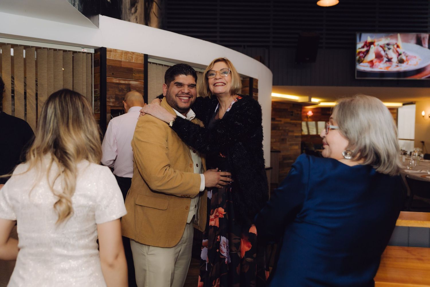 Groom in tan suit laughs with female guest in black dress at casual wedding reception venue.