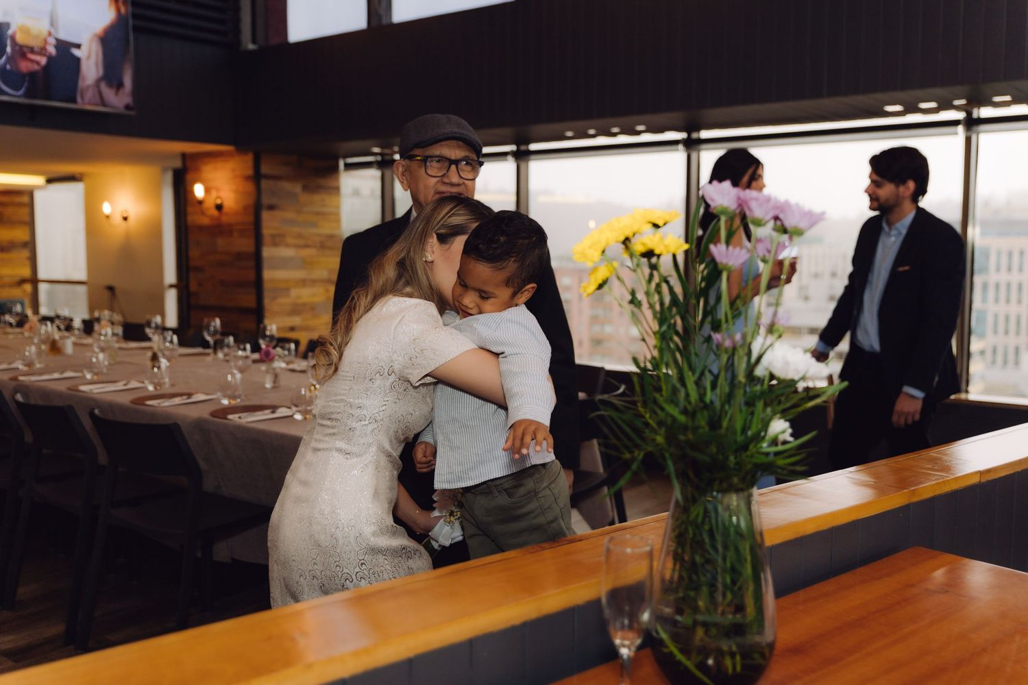 Bride in white dress hugs a child at wedding reception with floral centerpiece in foreground.