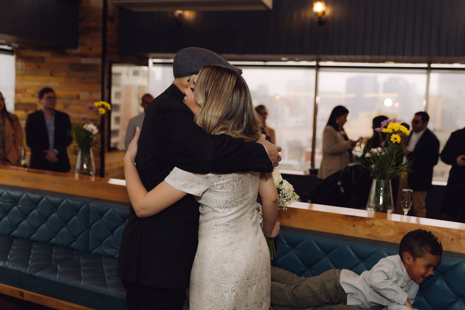 Bride in lace gown shares heartfelt hug with guest in cap at warmly lit wedding reception.