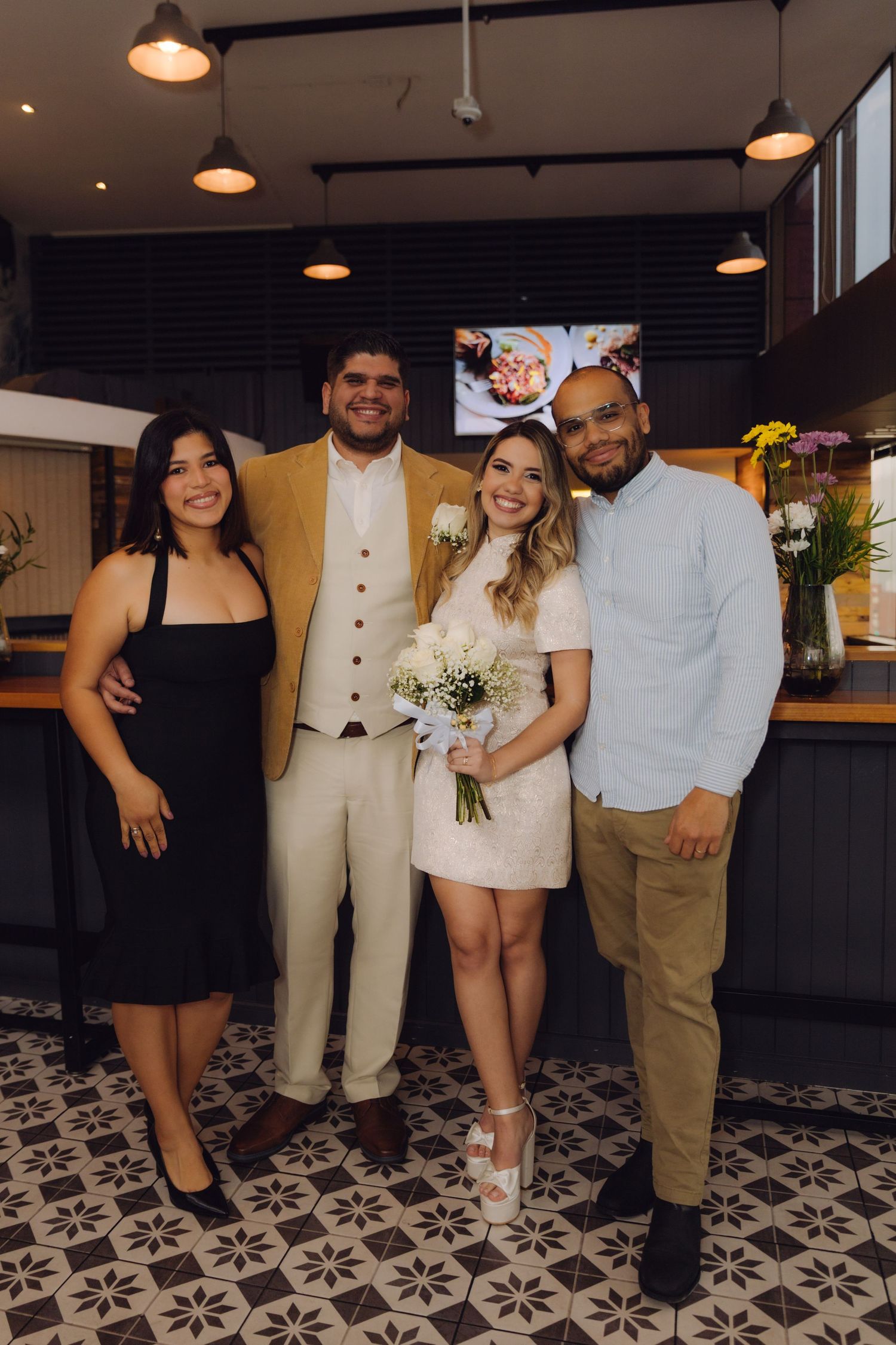 Wedding couple in white dress and gold suit poses with two guests at indoor celebration.