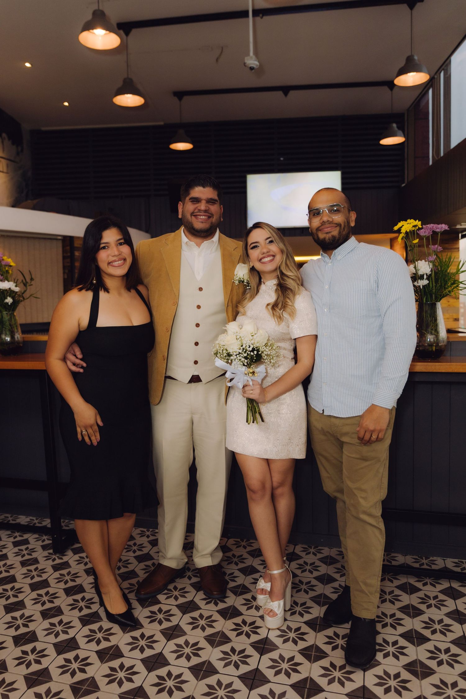 Smiling bride holding white bouquet stands with groom and two friends at wedding reception.