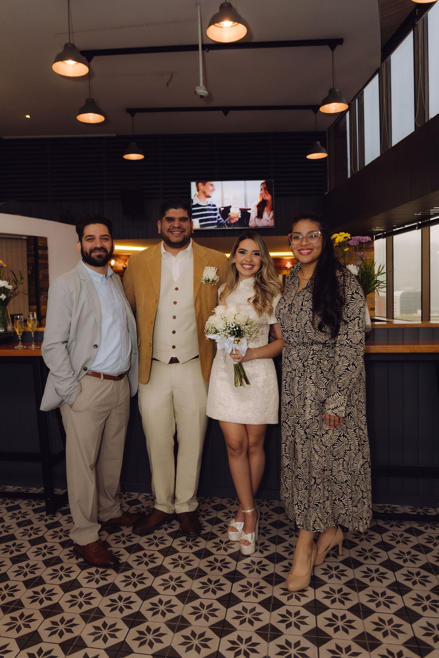 Bride holding white floral bouquet smiles with groom and two guests at wedding reception.