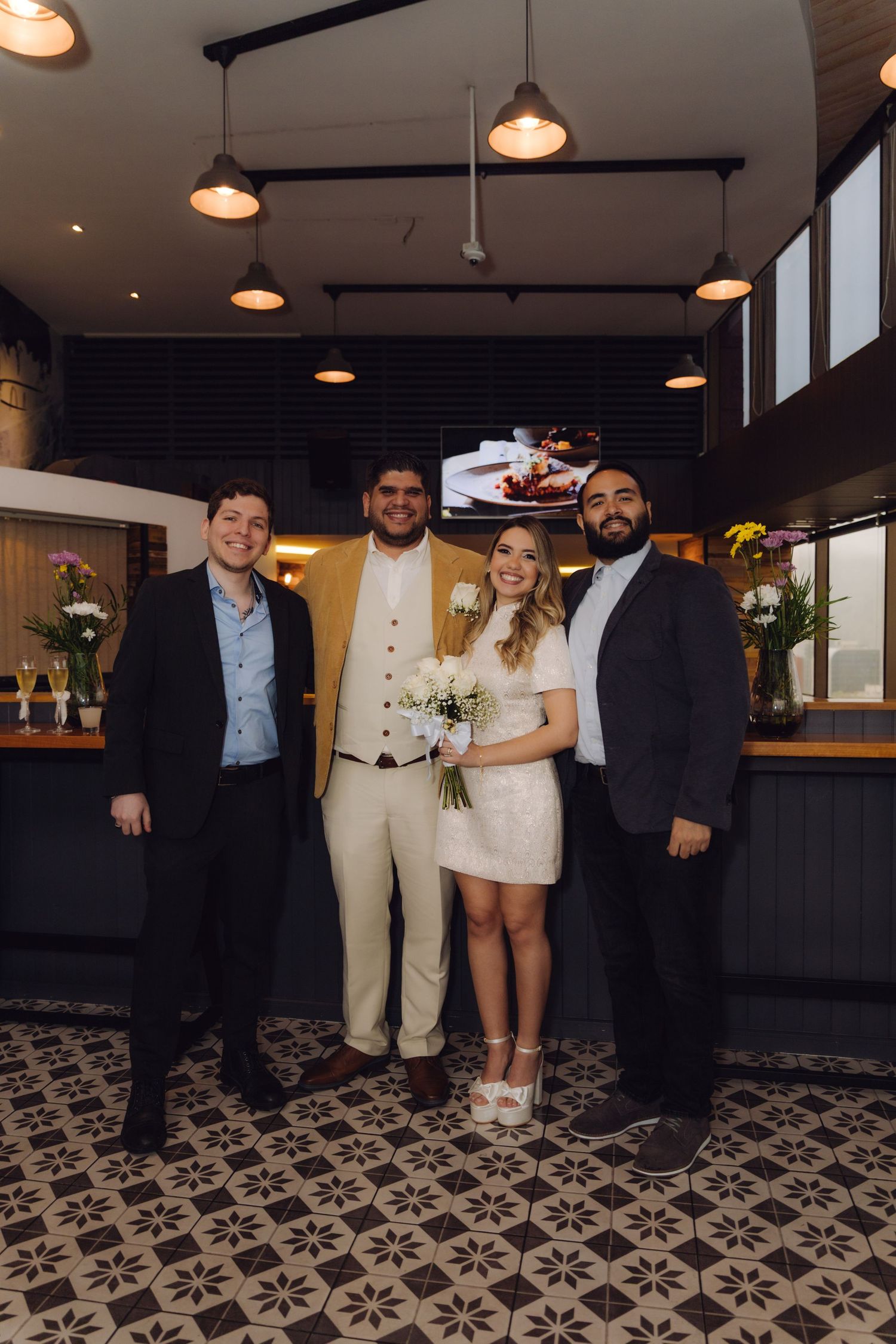 Groom in gold blazer and bride with bouquet pose with two suited guests at reception.