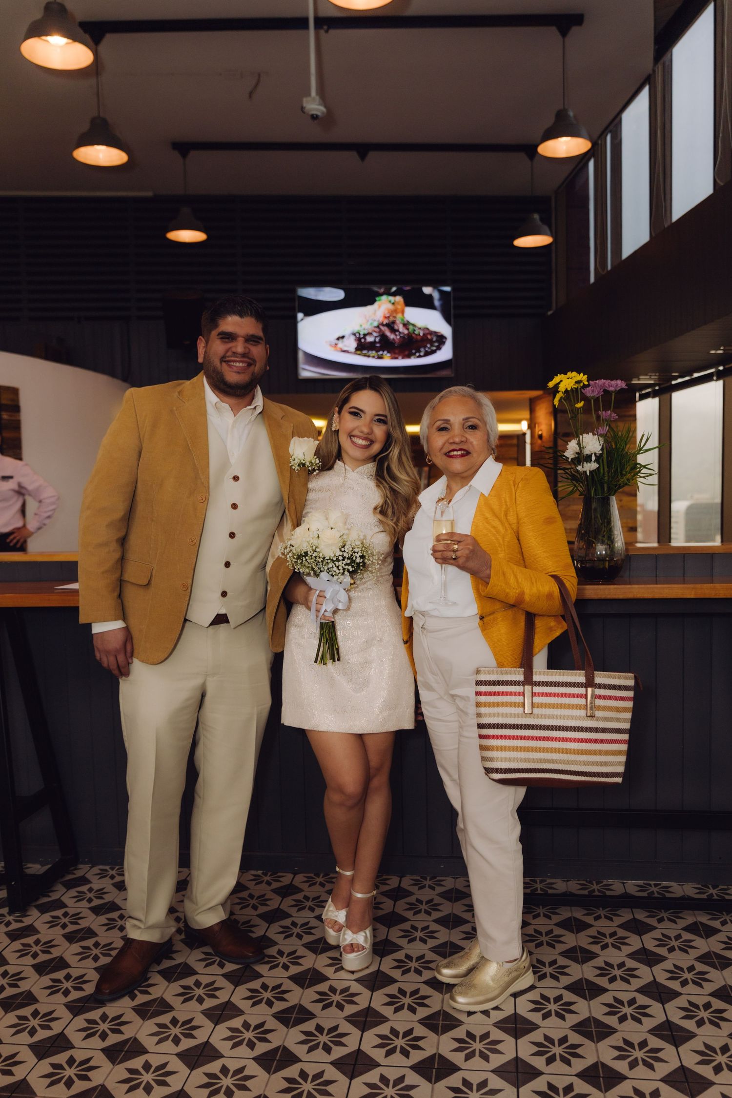 Bride in white mini dress and groom in gold jacket pose with older female guest.