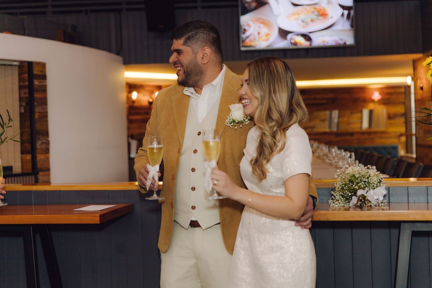 Newlyweds share a joyful moment holding champagne glasses at their wedding reception bar.