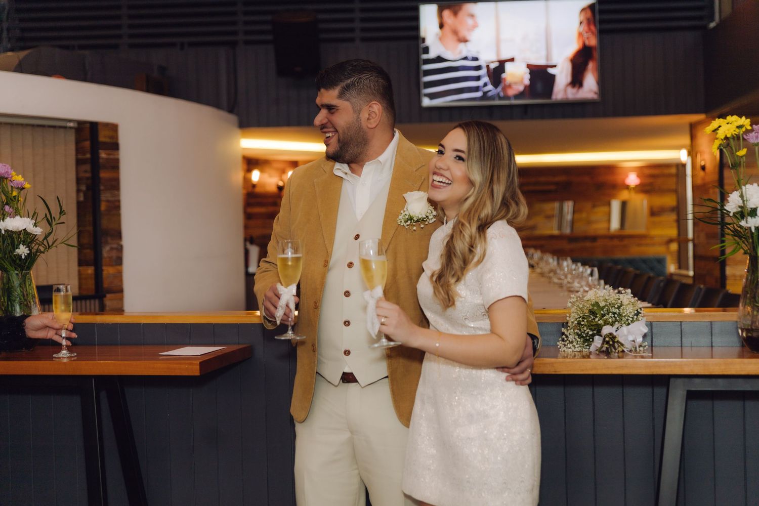Smiling newlywed couple holding champagne glasses at a bar, groom in mustard jacket and bride in white mini dress.