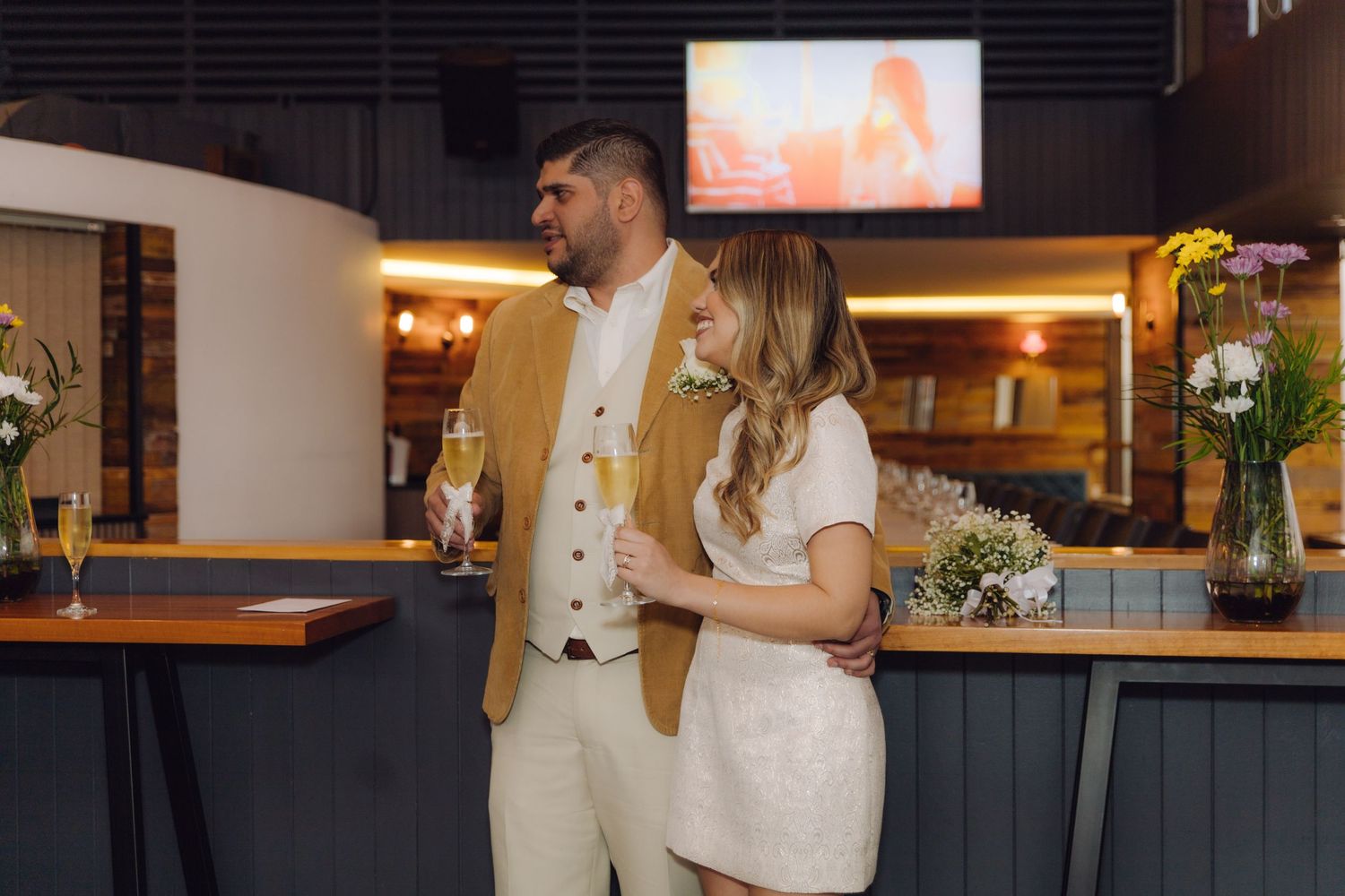 Newlywed couple standing at a bar, groom in mustard blazer embracing bride in white dress holding a bouquet.