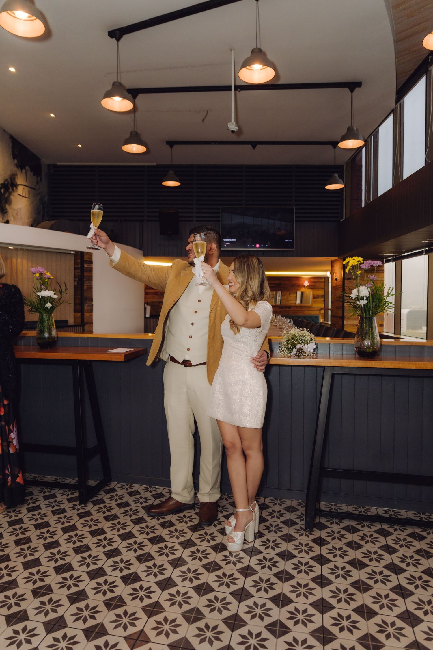 Newlywed couple raising champagne glasses in a toast inside a stylish bar venue with patterned tile floors.