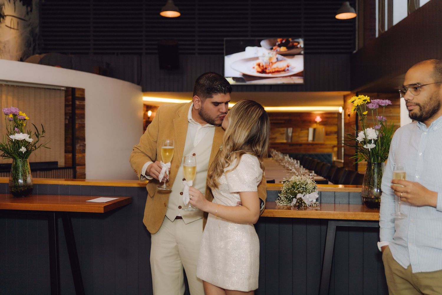 Newlywed couple sharing a kiss at a bar while a guest stands nearby holding a champagne glass.