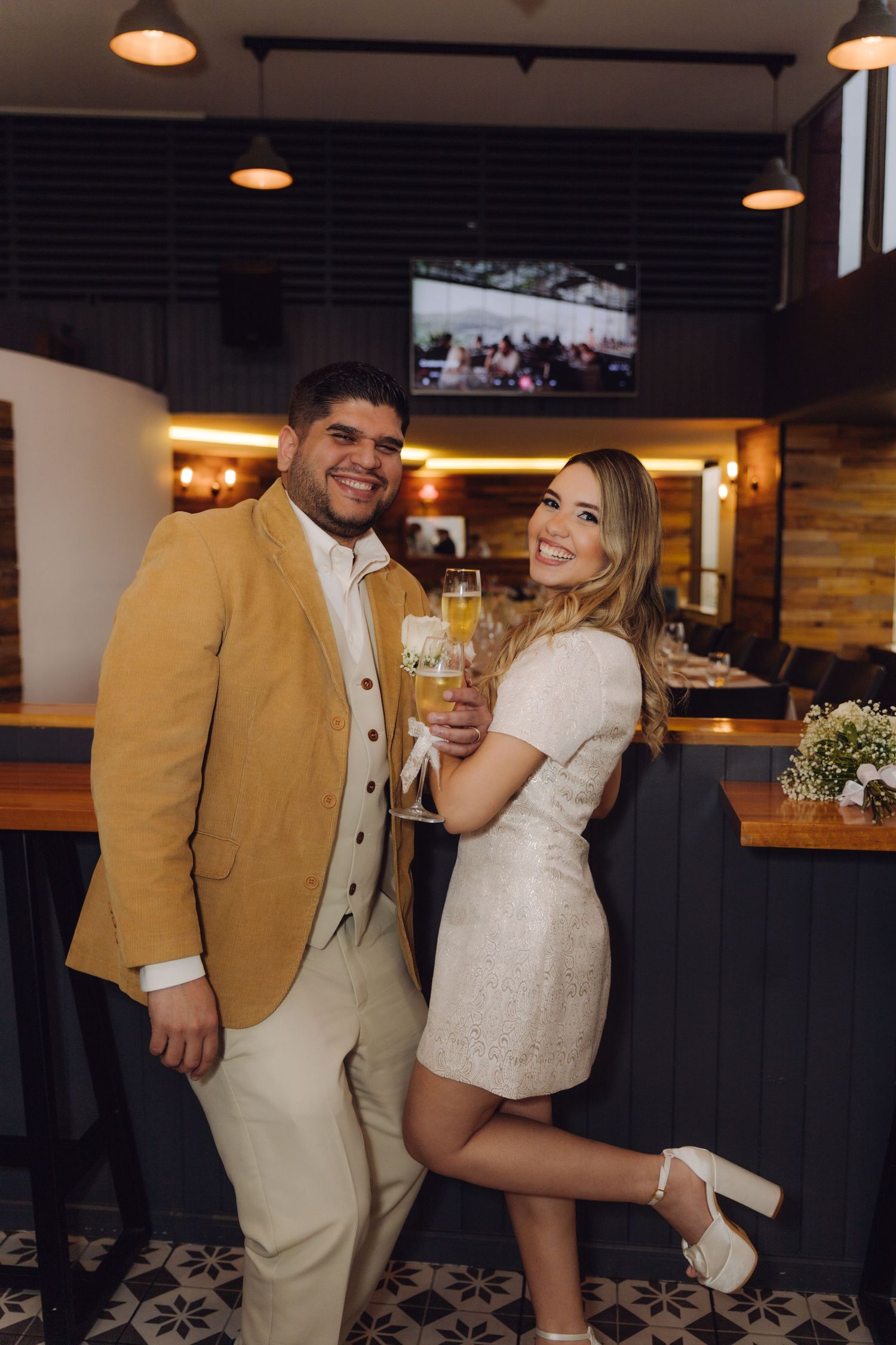 Happy newlywed couple smiling and clinking champagne glasses at a bar, bride in white mini dress and groom in mustard jacket.
