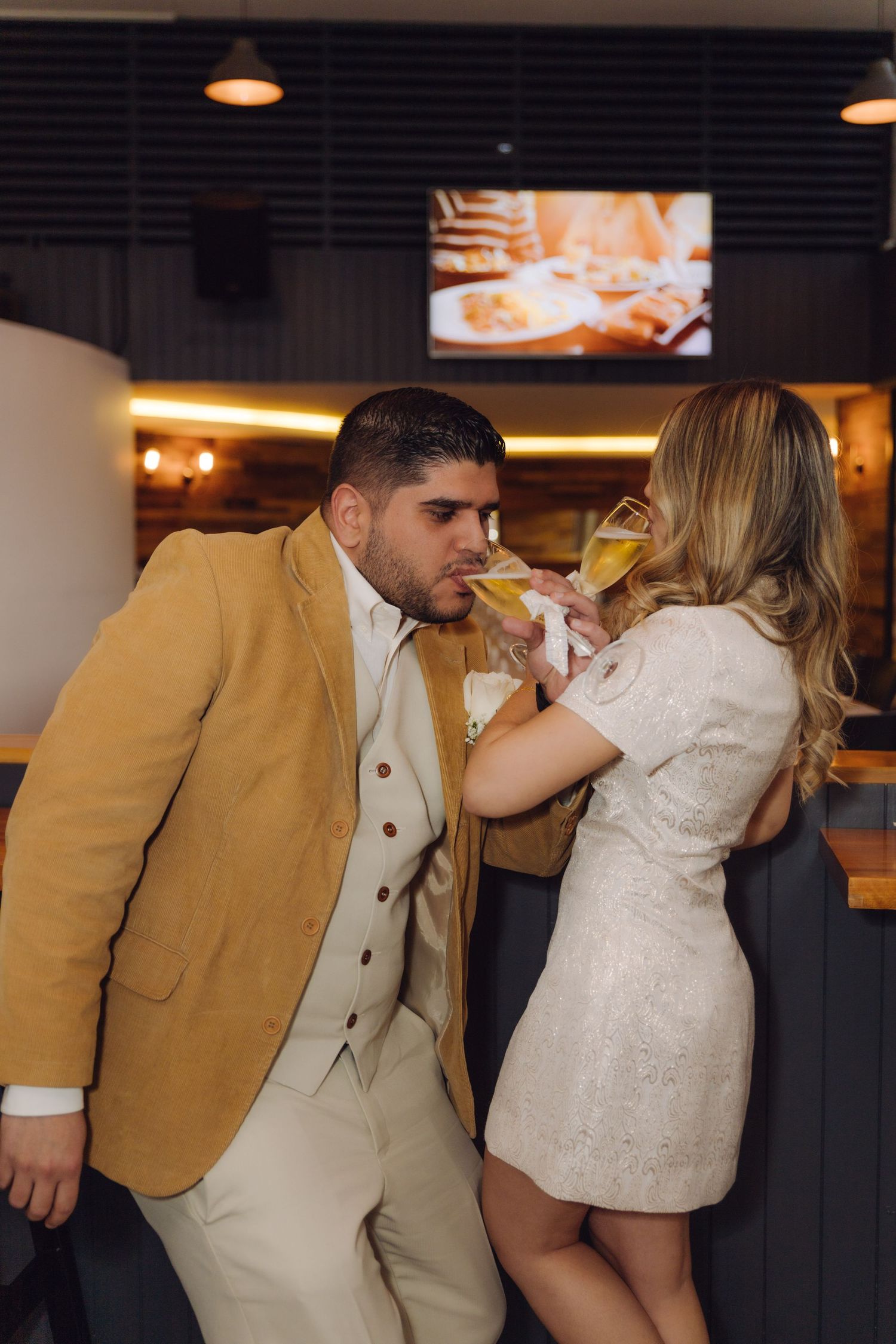 Newlywed couple drinking champagne together at a bar, groom in mustard blazer and bride in white lace dress.