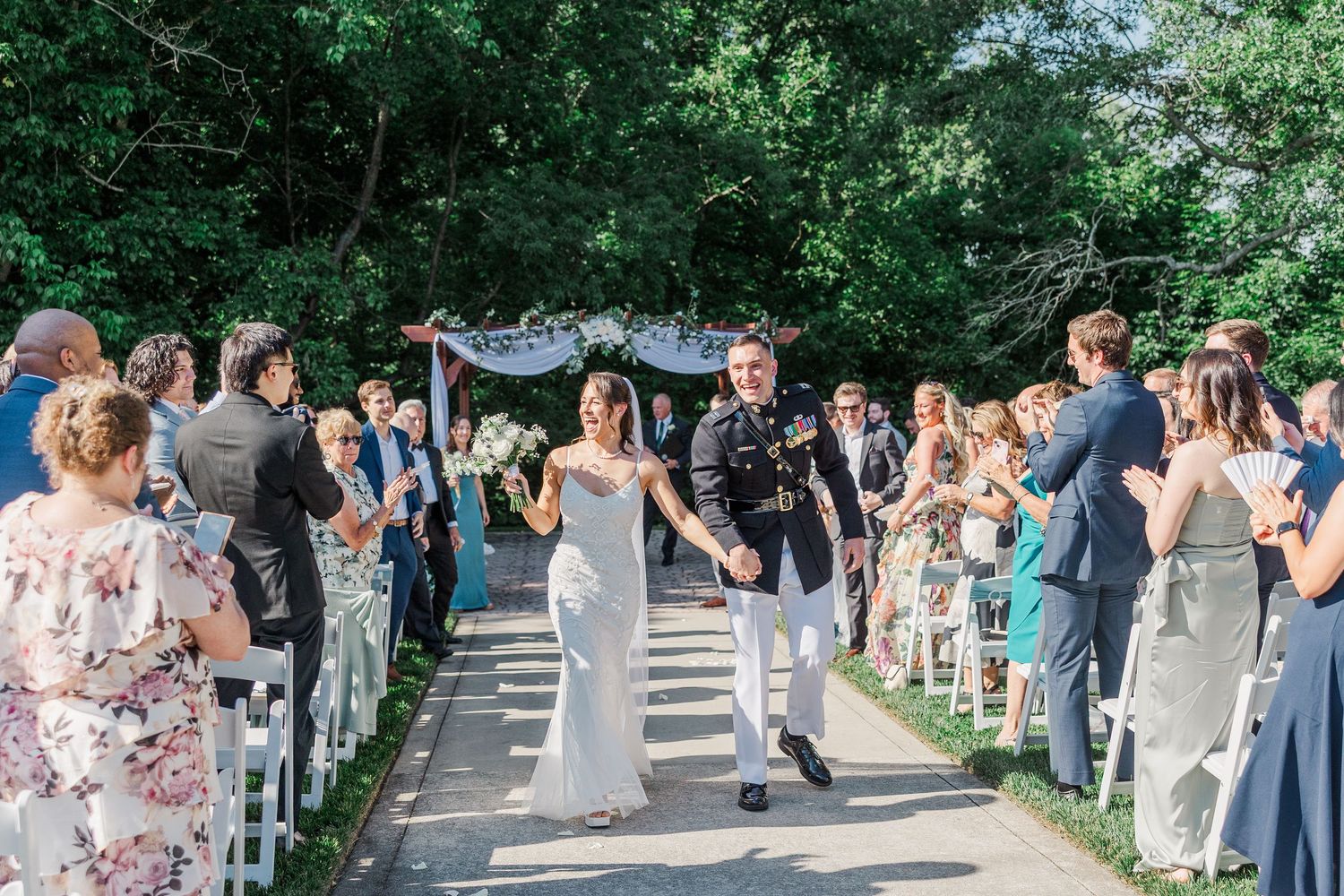 Bride and groom walk down outdoor aisle after wedding ceremony, surrounded by cheering guests under lush green trees.