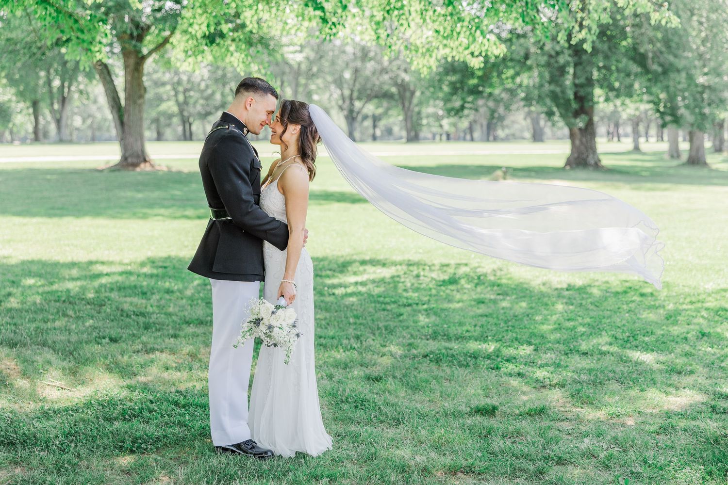 A bride and groom share a romantic kiss in a lush green park, her veil flowing gracefully in the breeze.