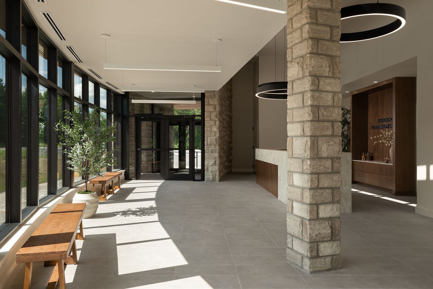 Modern building lobby with stone columns, floor-to-ceiling windows, wooden benches, and circular pendant lights on white ceiling.