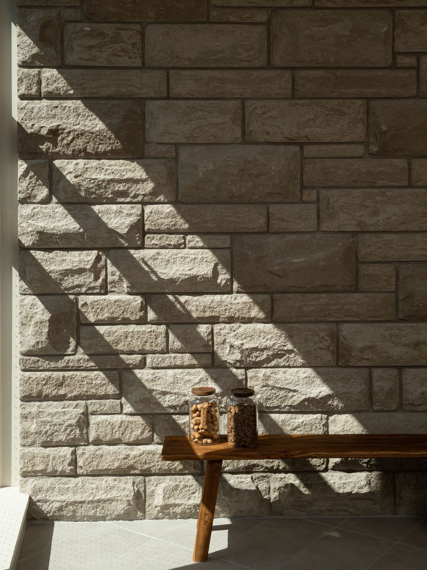 Wooden bench with a glass jar against a sunlit rustic stone wall casting dramatic diagonal shadows.