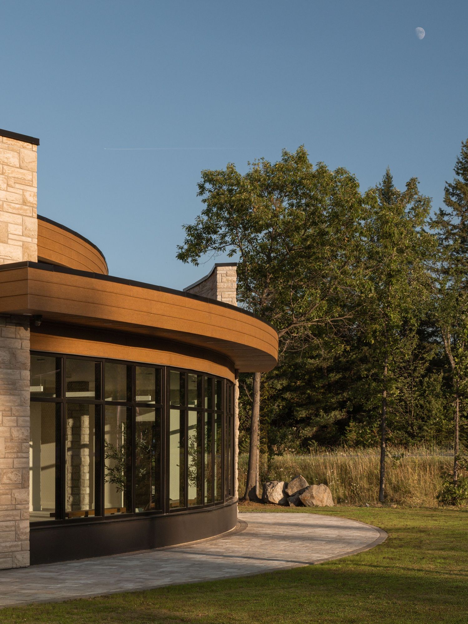Curved modern building with wood soffit, stone facade, and floor-to-ceiling windows set against lush trees and blue sky.